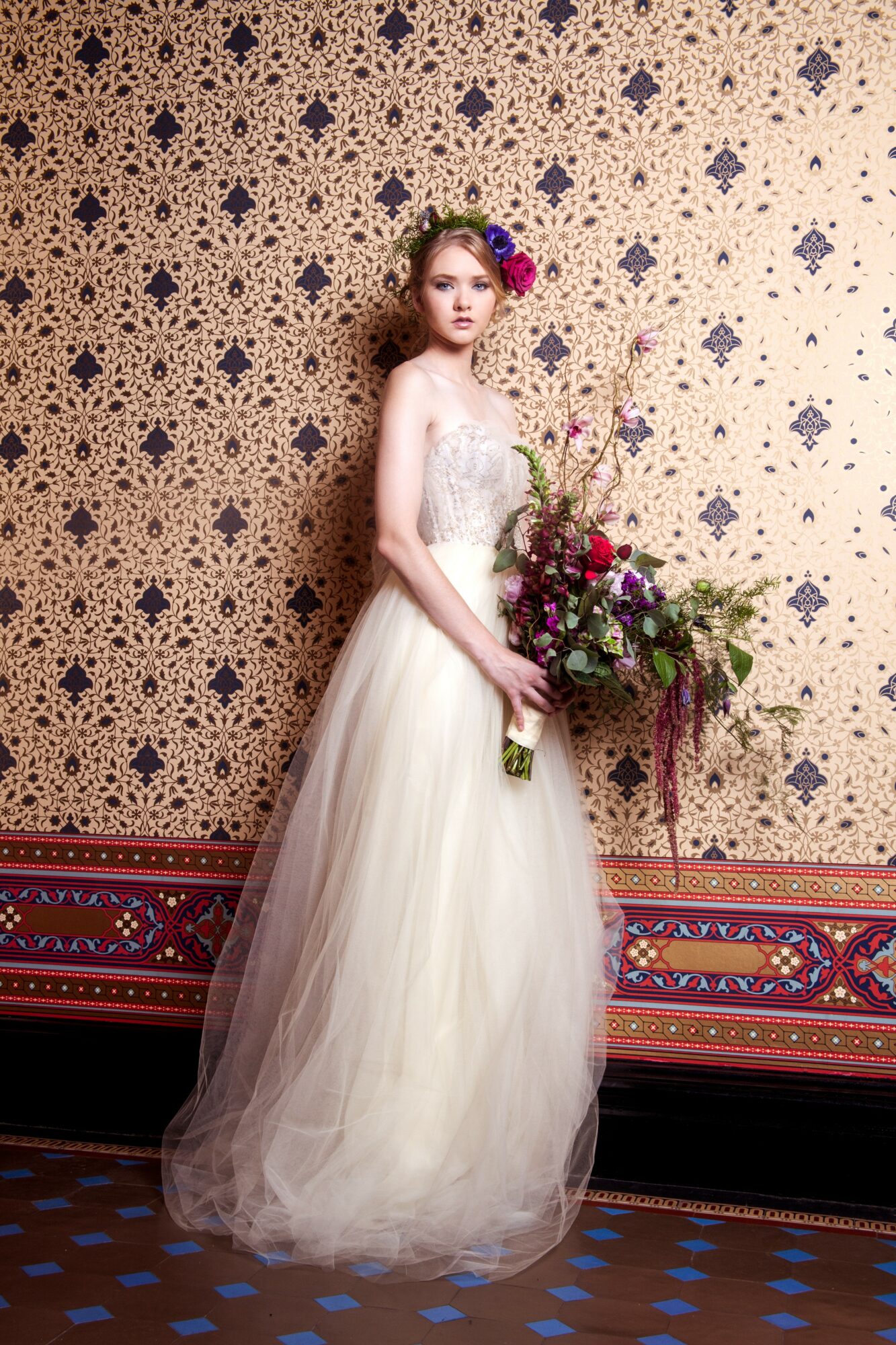 Young woman in a white dress holding a bouquet, standing against a patterned wall with a decorative carpeted floor.