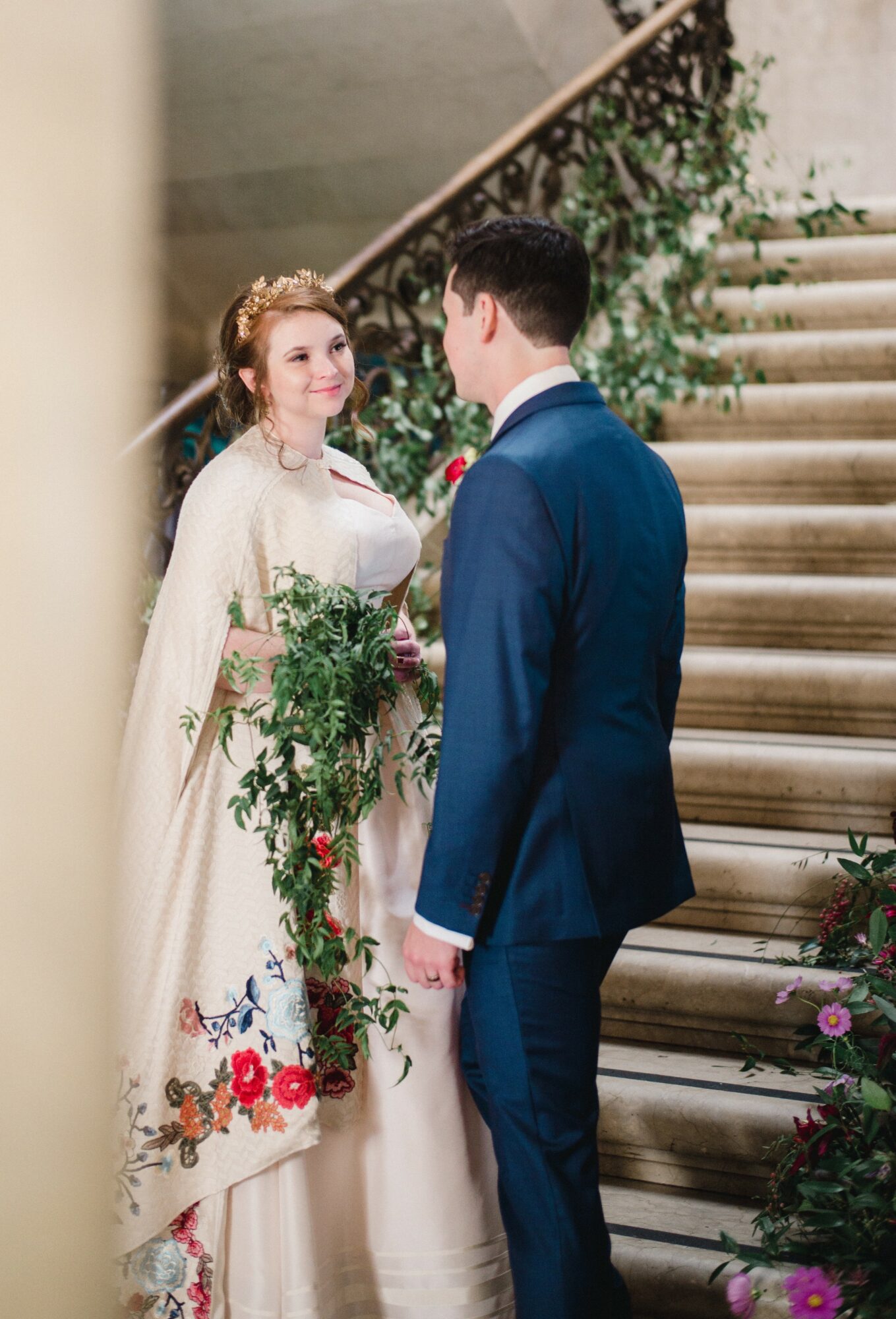Bride and groom stand on staircase, facing each other, bride holding a bouquet, surrounded by greenery and flowers.