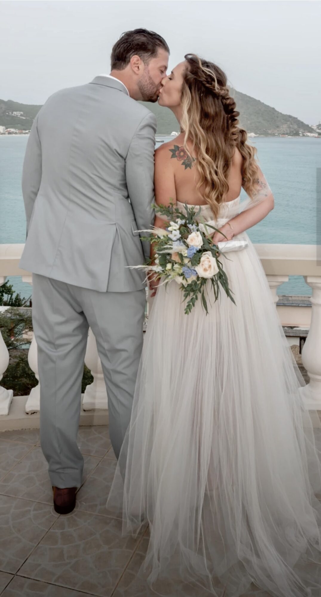A couple in wedding attire kissing outdoors with a body of water and mountains in the background.