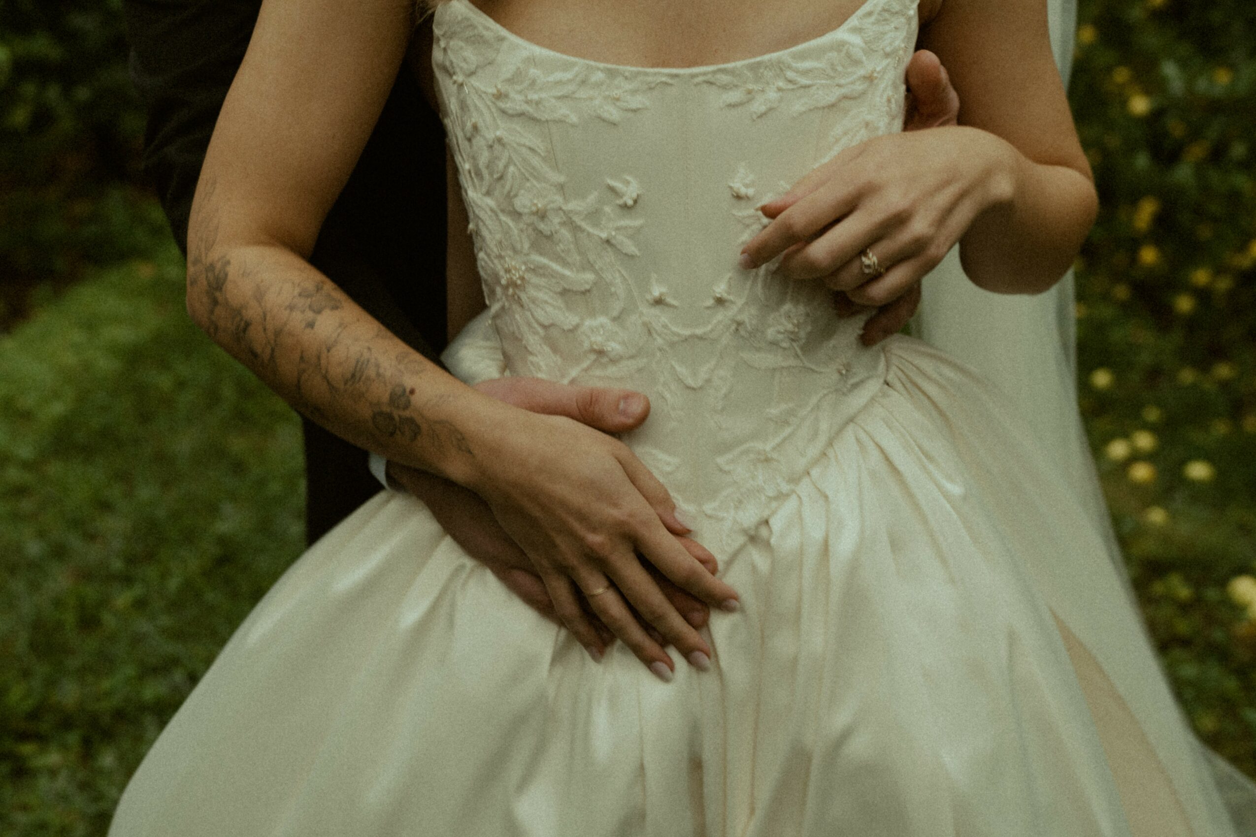 Close-up of a person in a white dress with hands resting on their waist, outdoors with greenery in background.