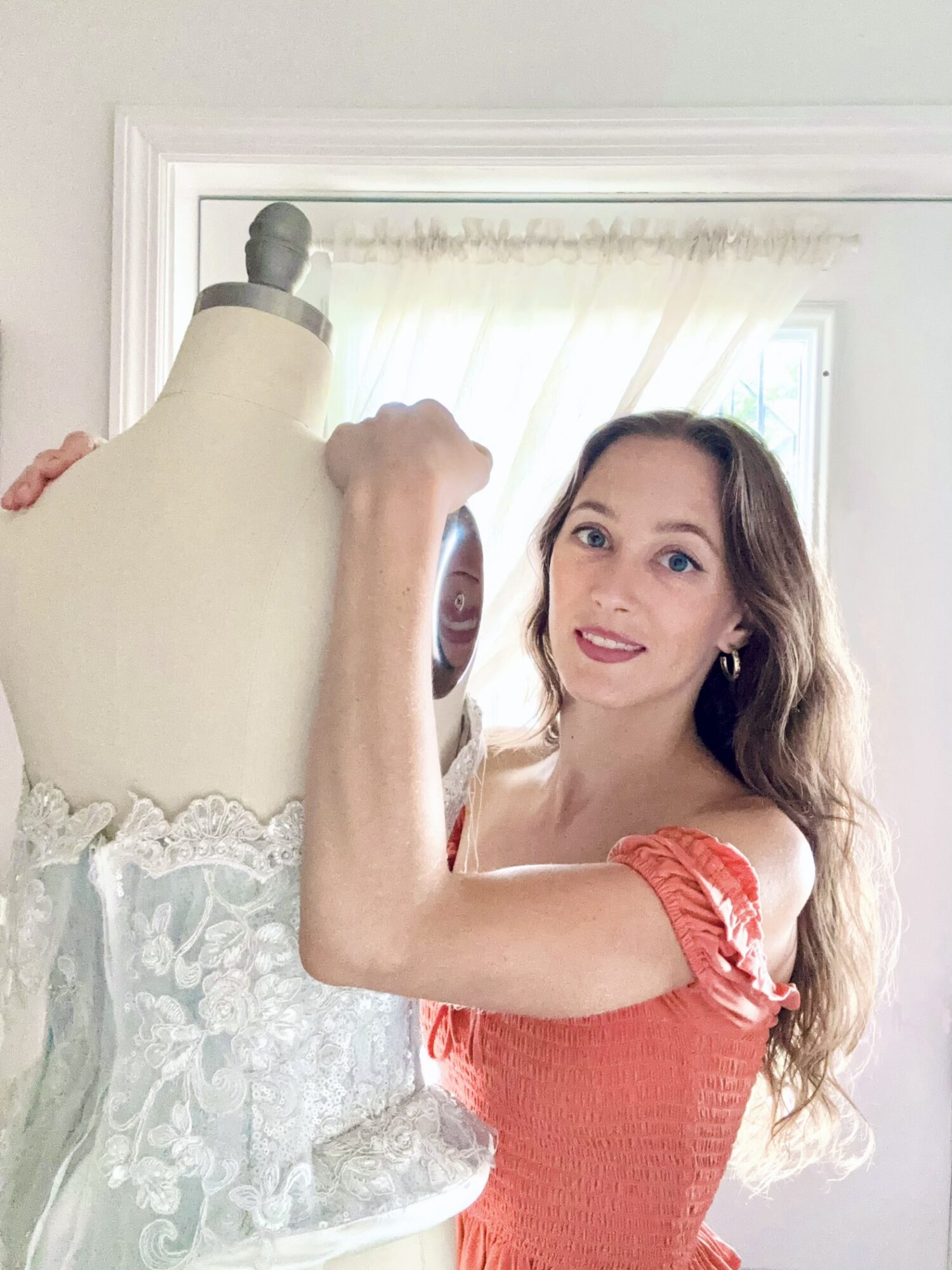 Young woman with long hair smiling, wearing a red dress, standing next to a dress form with a lace garment, in a bright room.