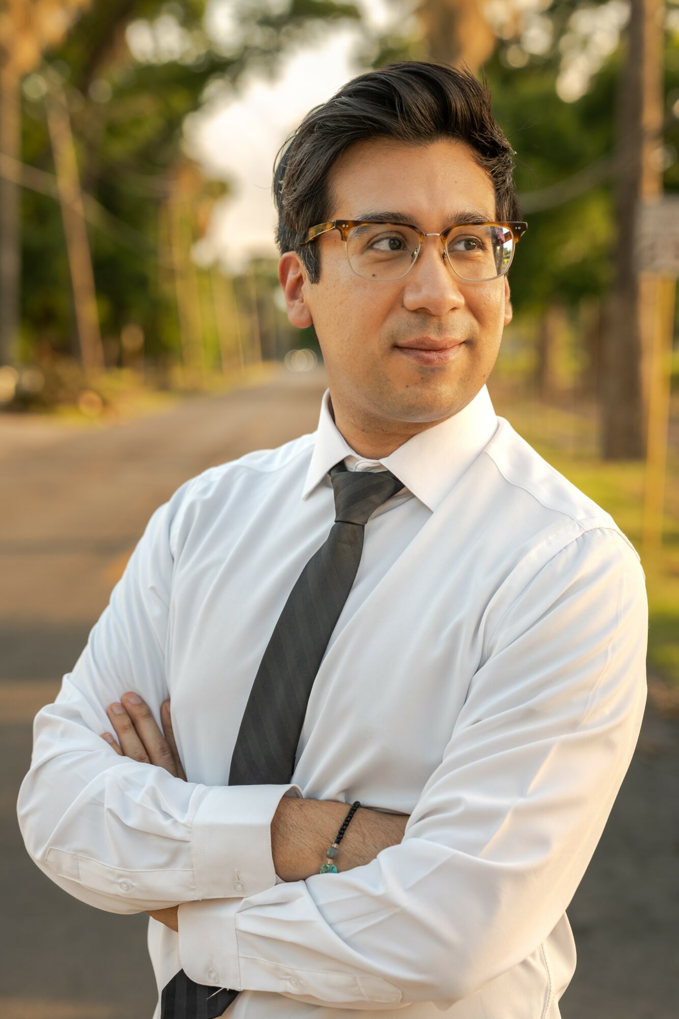 Man with glasses in white shirt and black tie standing outdoors with trees in background