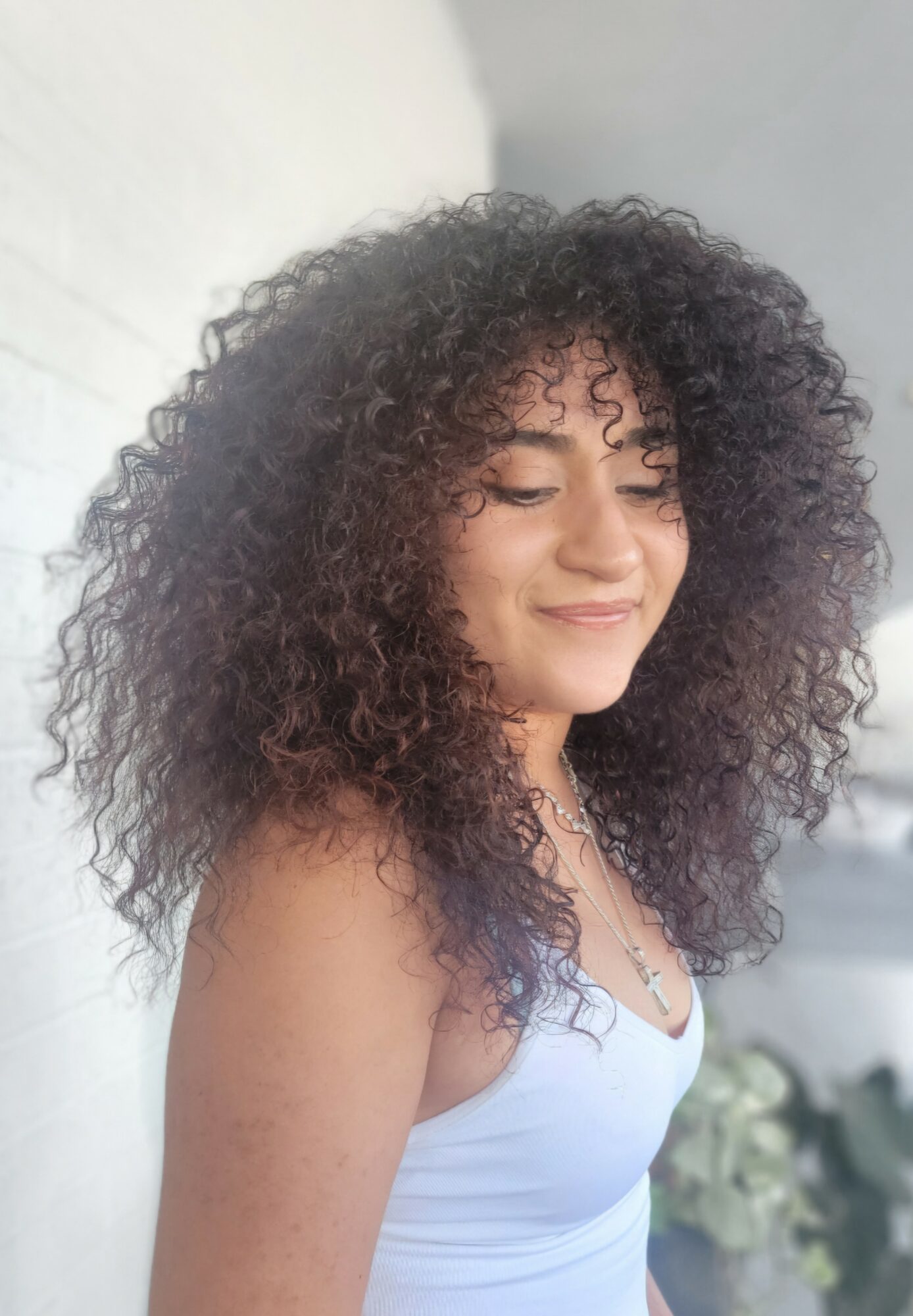 Young woman with curly hair smiling, wearing a white top, facing slightly to the right, against a light background.
