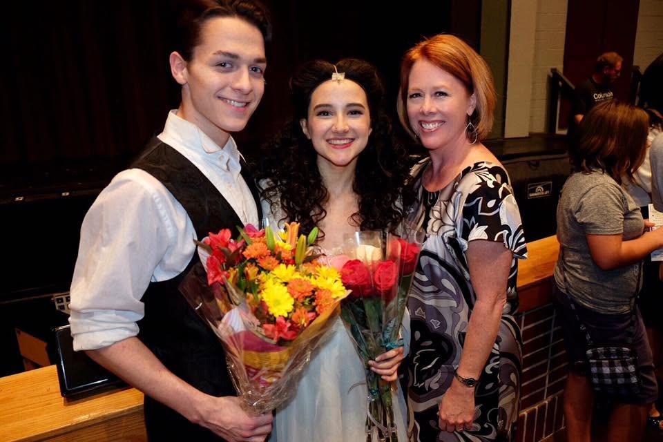 Three people smiling, two women and one man, holding flowers, in an indoor setting with a stage in the background.