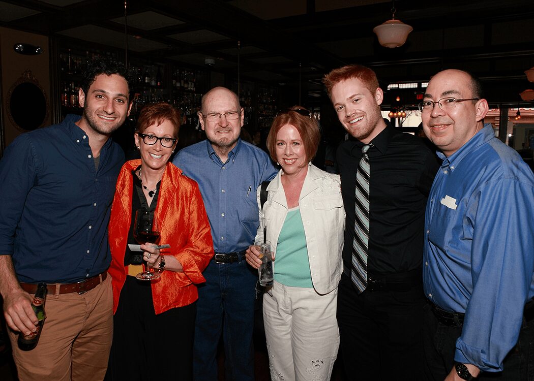 Group of six people smiling, standing together in a dimly lit indoor setting, some holding drinks, dressed in casual and semi-formal attire.