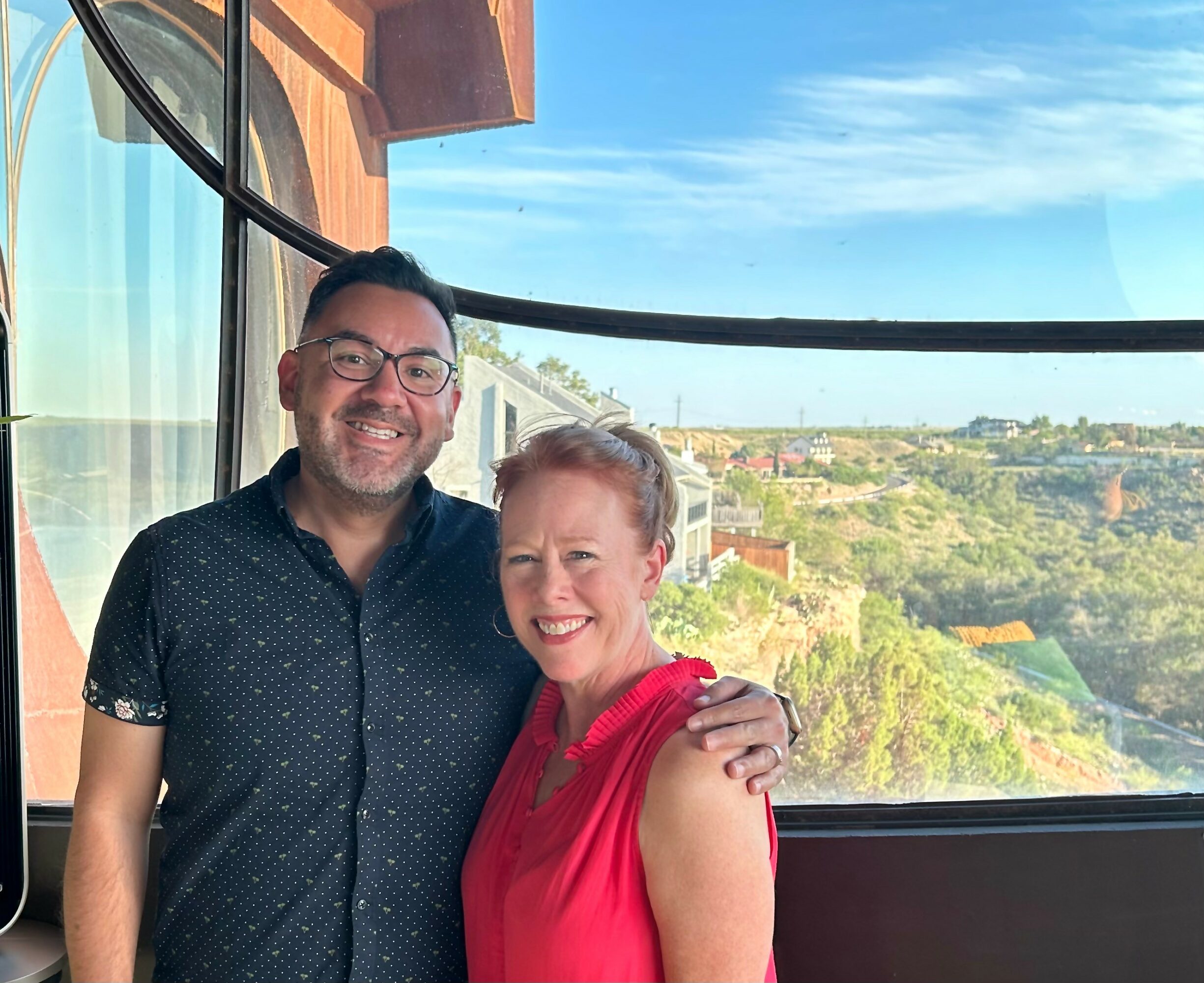 Two people smiling indoors with a large window showing a landscape view, one with glasses and a dark shirt, the other in a red top.