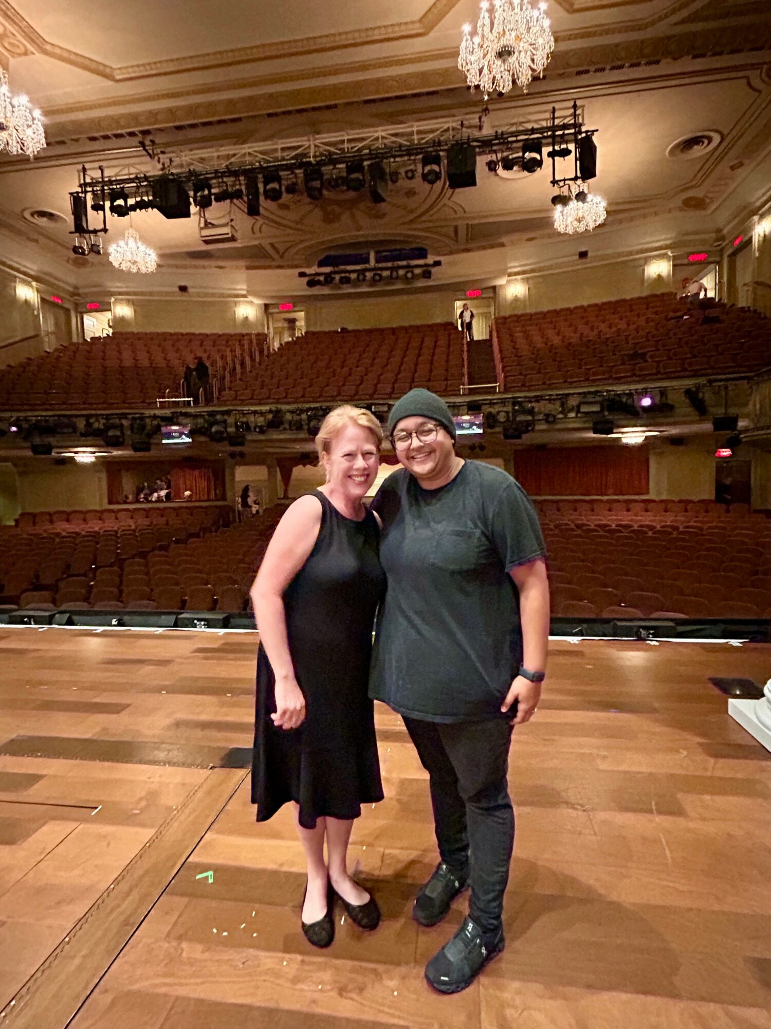 Two people standing on a stage in an empty theater, smiling, with chandeliers and seating in the background.