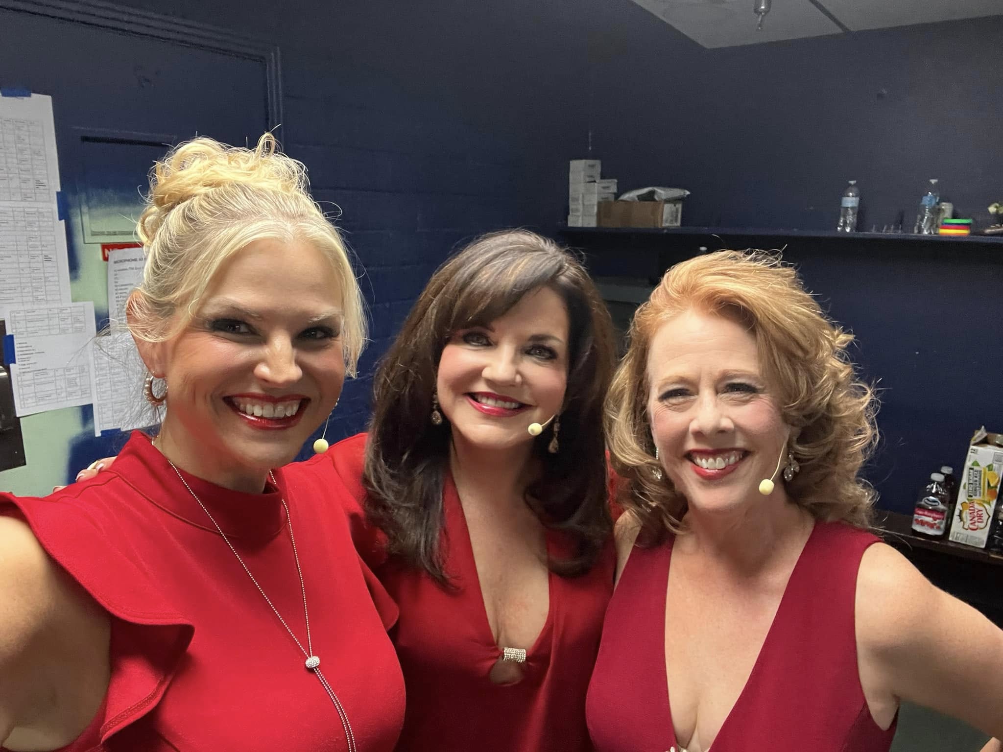 Three women smiling, wearing red tops, standing indoors with dark background and shelves, close together.