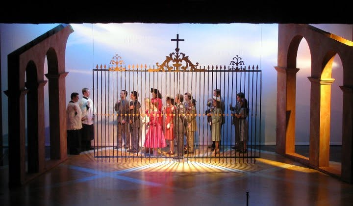 Group of people standing behind a decorative gate with a cross, framed by arches, in a courtyard setting.