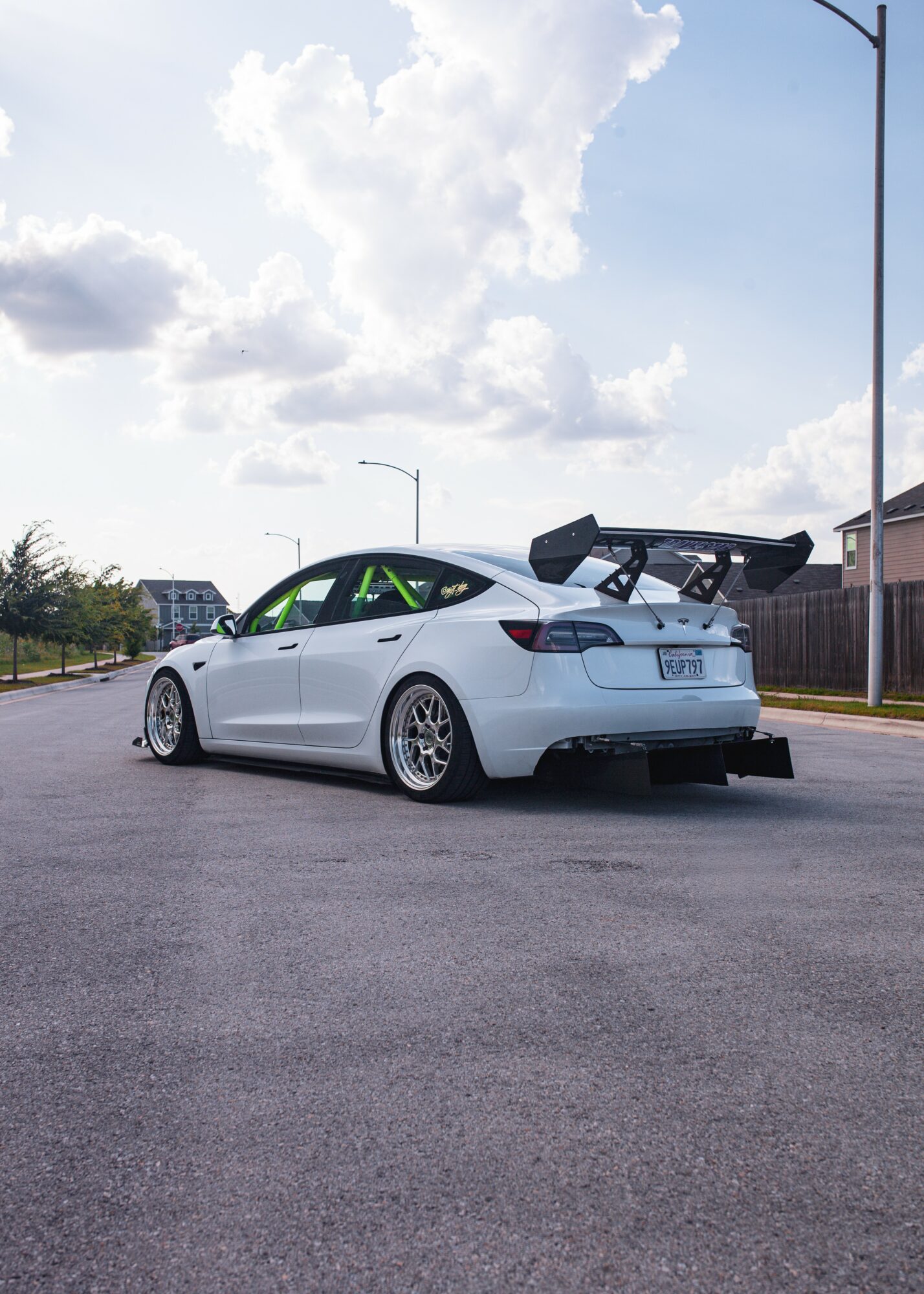 White sports car with a large rear wing parked on a street under a partly cloudy sky.