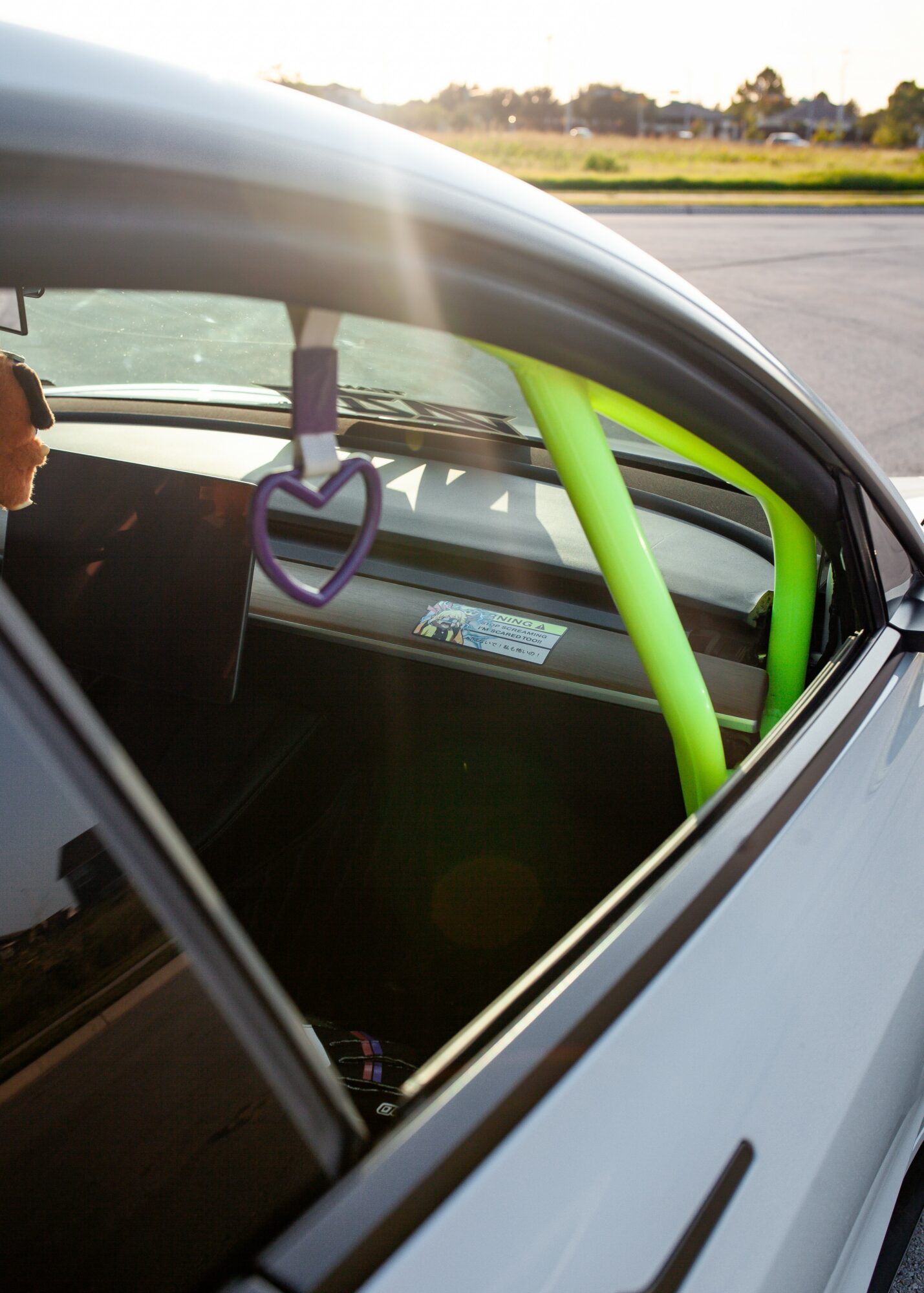 View through car window showing a green roll cage inside the vehicle, with a purple heart-shaped hanging ornament, and a dashboard.