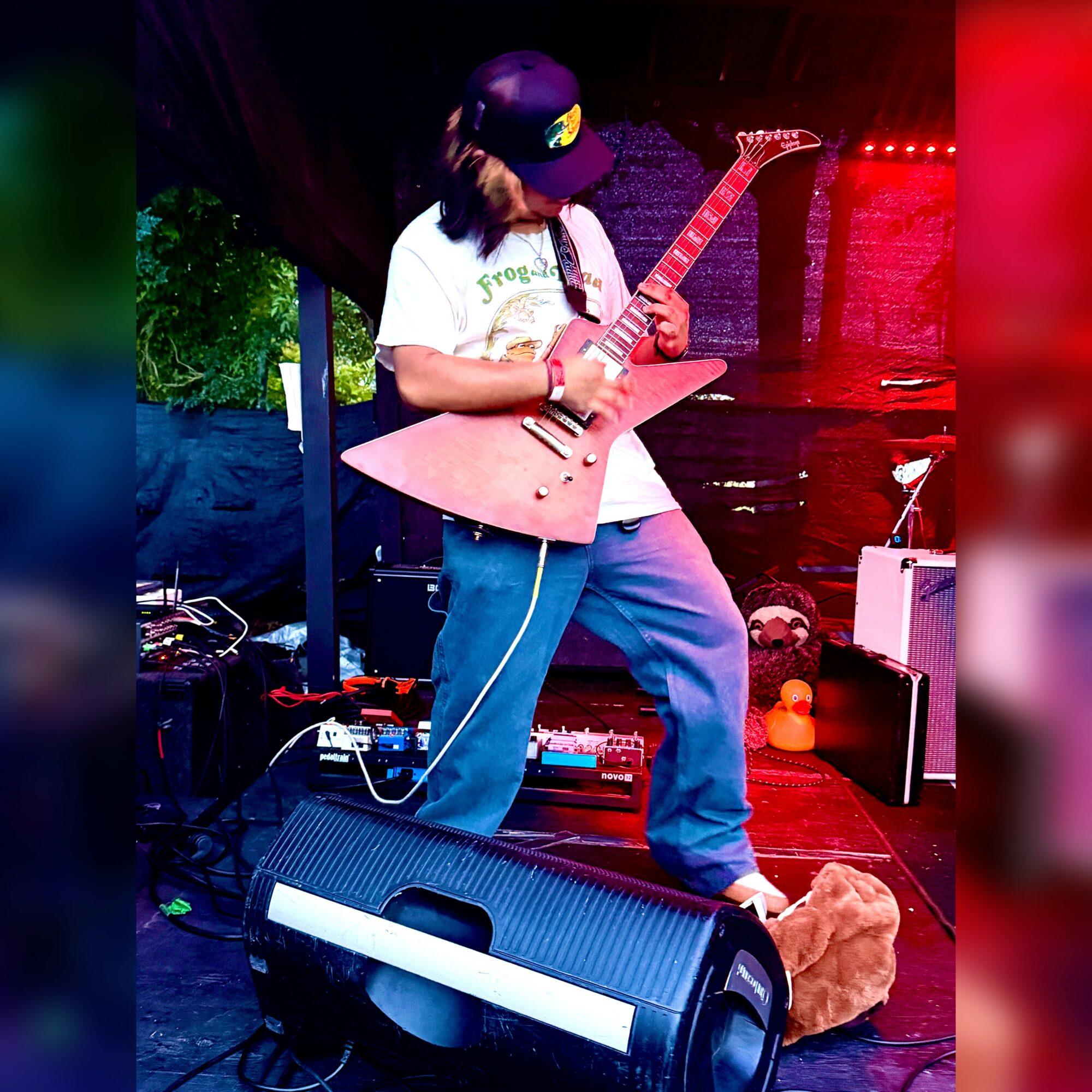Person playing an electric guitar on stage, wearing a cap and casual clothes, with musical equipment around.