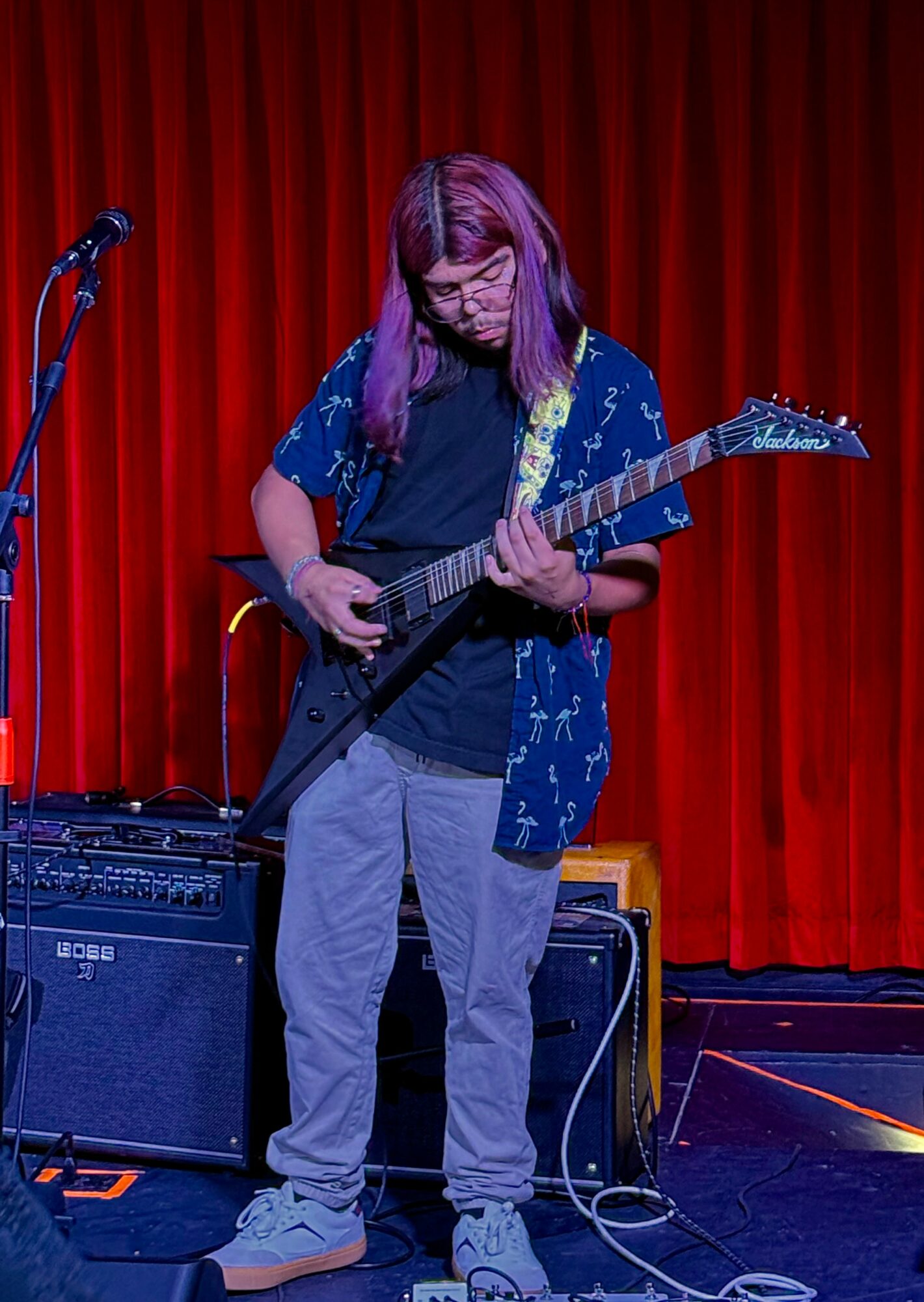 Person with long hair playing an electric guitar on stage with red curtains in the background.
