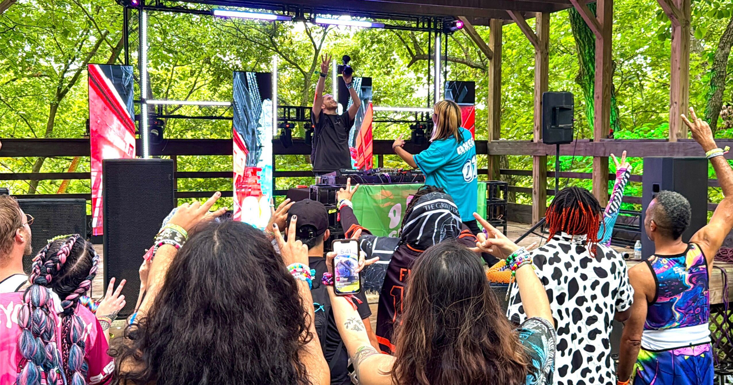 Group of people gathered outdoors watching a performance on stage with trees in background.