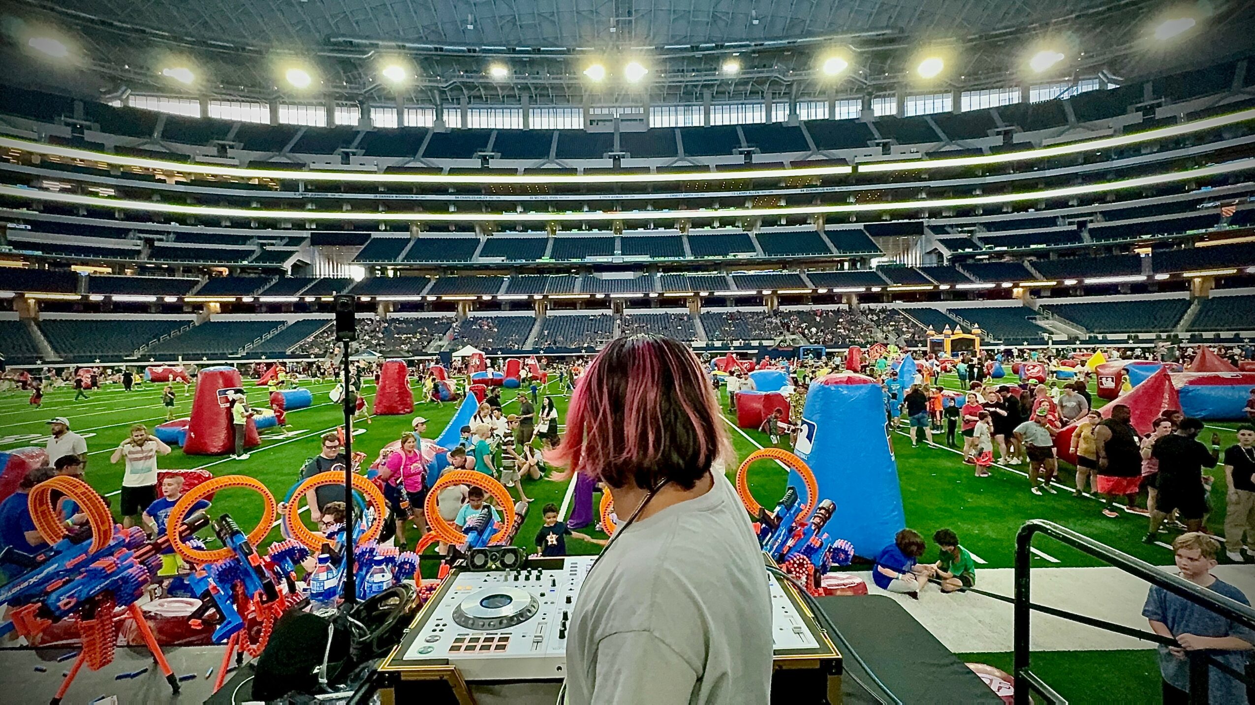 Person with pink hair at DJ booth overlooking a crowded stadium with colorful tents and people on the field.