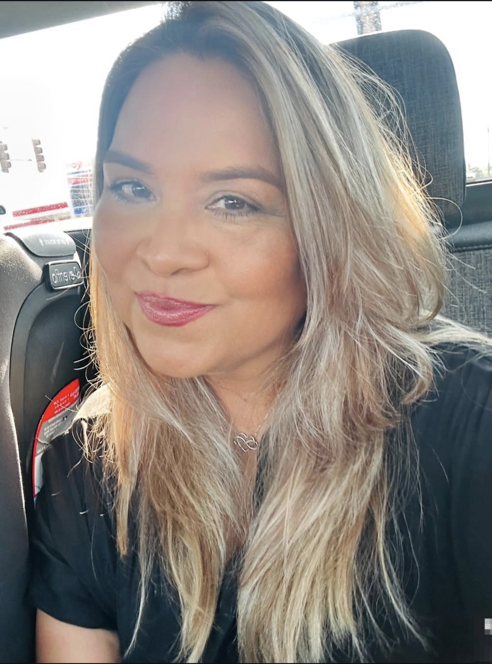 Woman with long, wavy hair smiling in a car, sunlight on her face, wearing a black top.