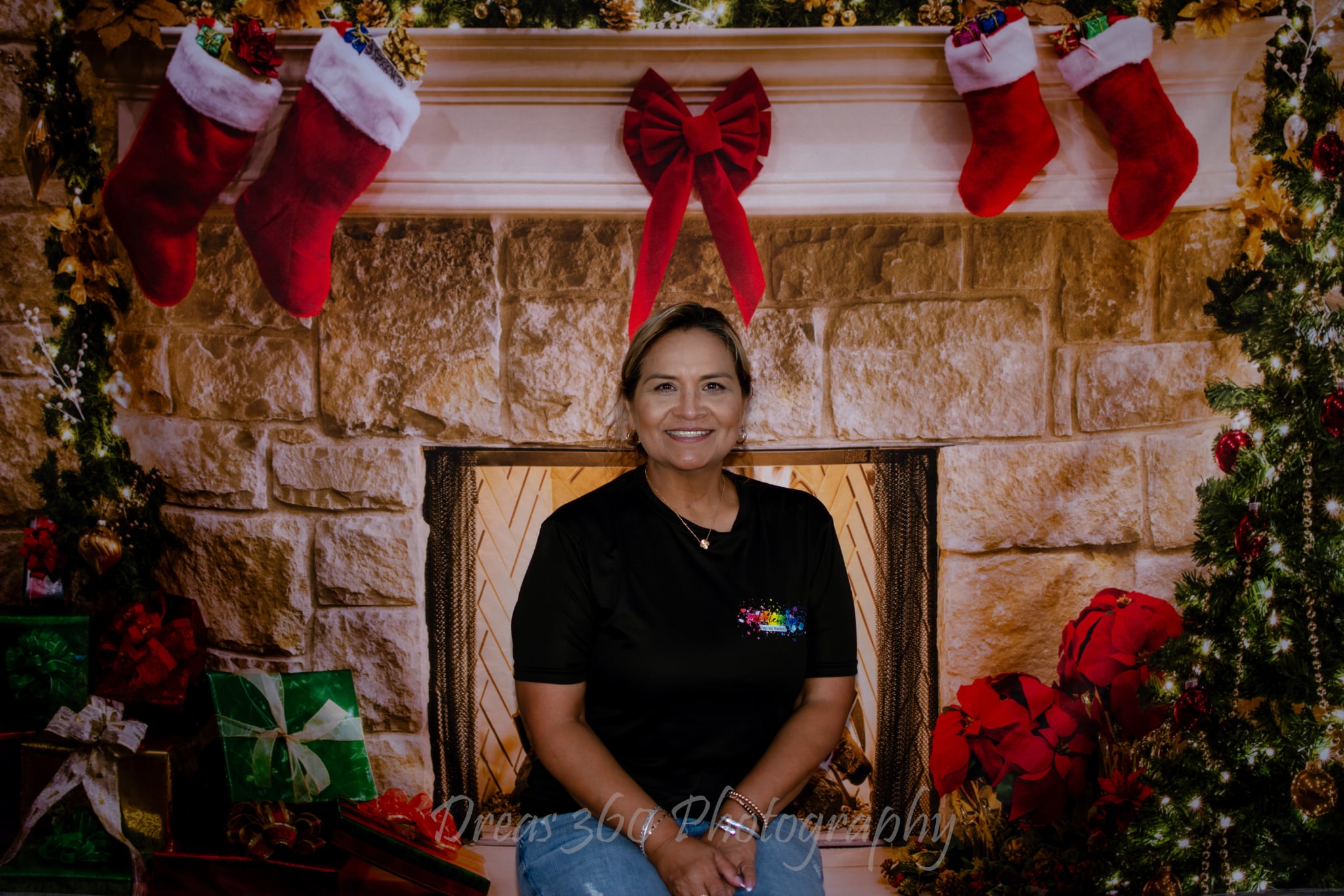 Woman sitting in front of a decorated fireplace with stockings and Christmas decorations, smiling, holiday setting.
