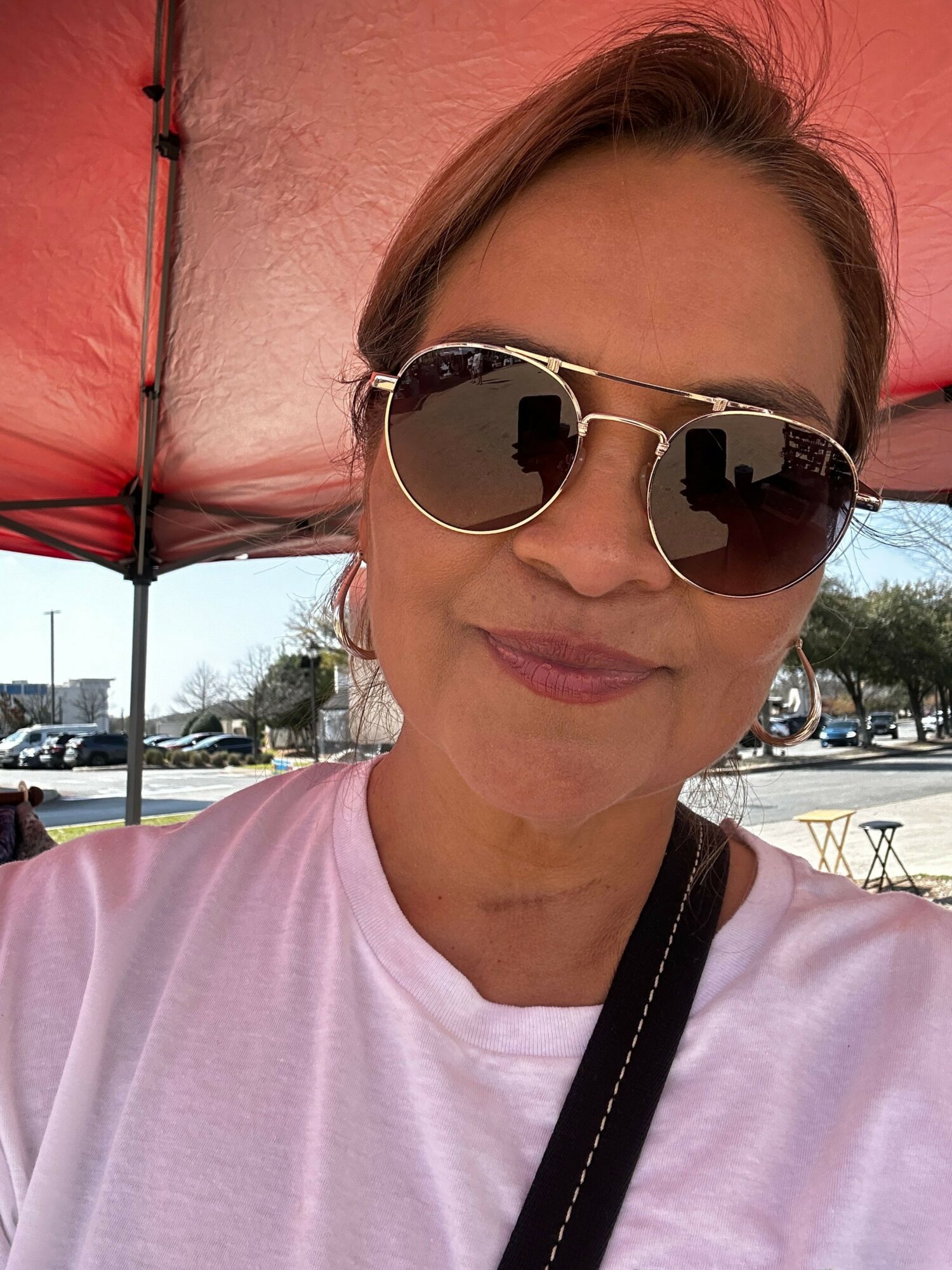 Woman wearing sunglasses and a white shirt, sitting outdoors with a pink wall behind her.