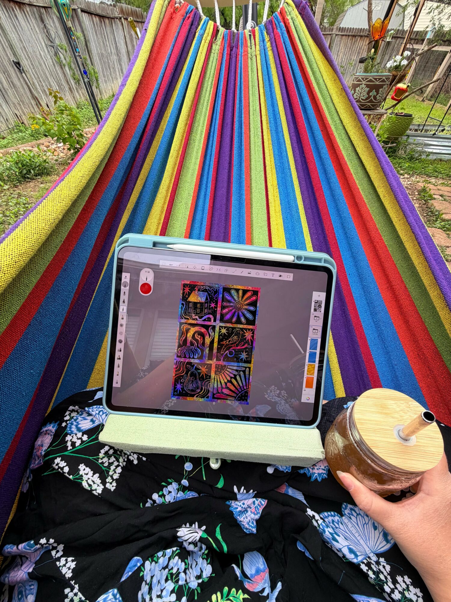 Person lying on a colorful hammock using a tablet, with a hand holding a wooden cup, outdoors in a garden.