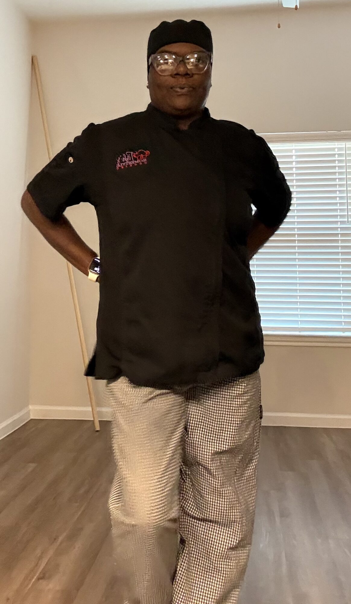 Person standing indoors with hands on hips, wearing a black chef's jacket and light-colored pants, in front of a window with blinds.