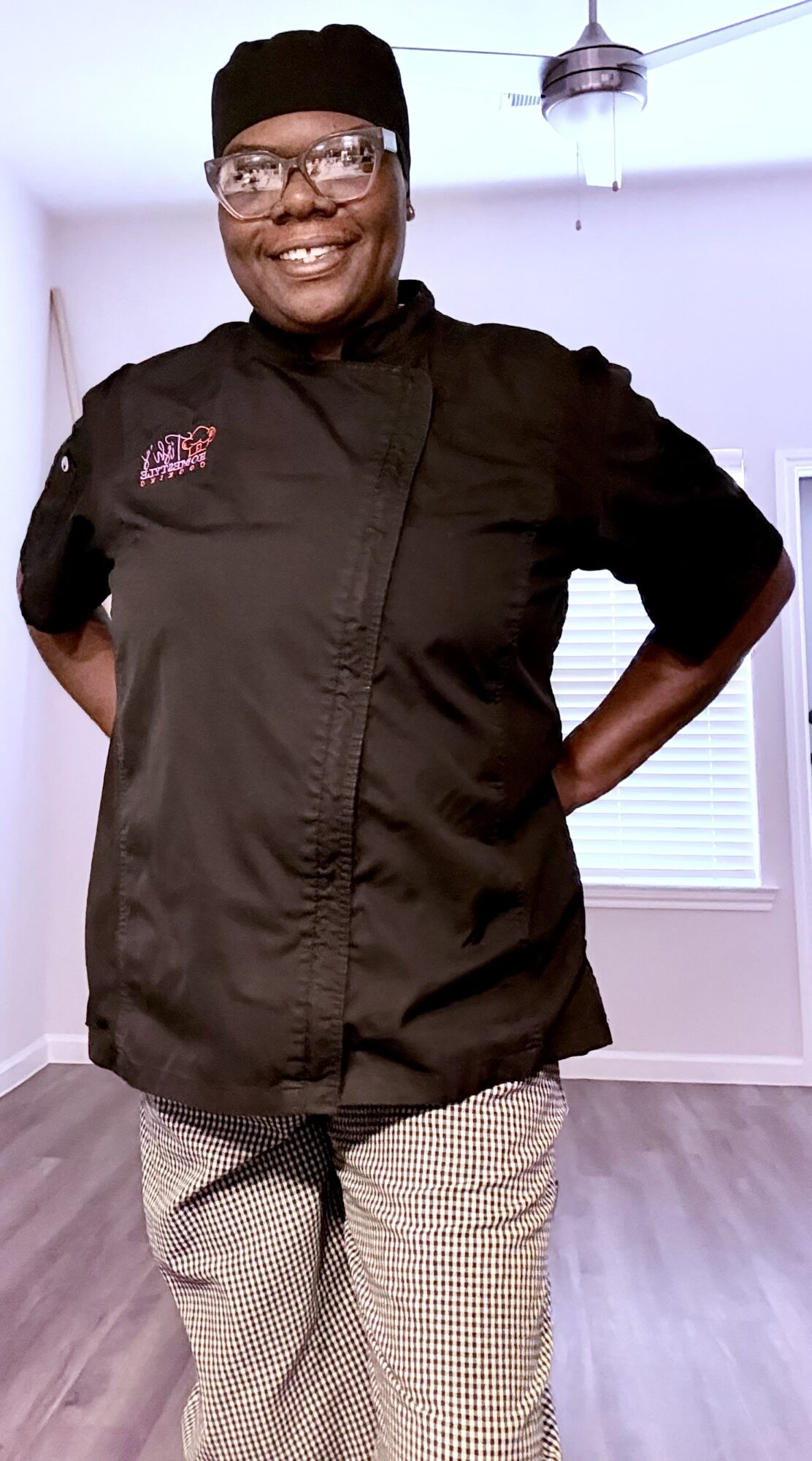 Person in chef uniform smiling with hands behind back indoors, ceiling fan visible, light-colored walls, and window with blinds.