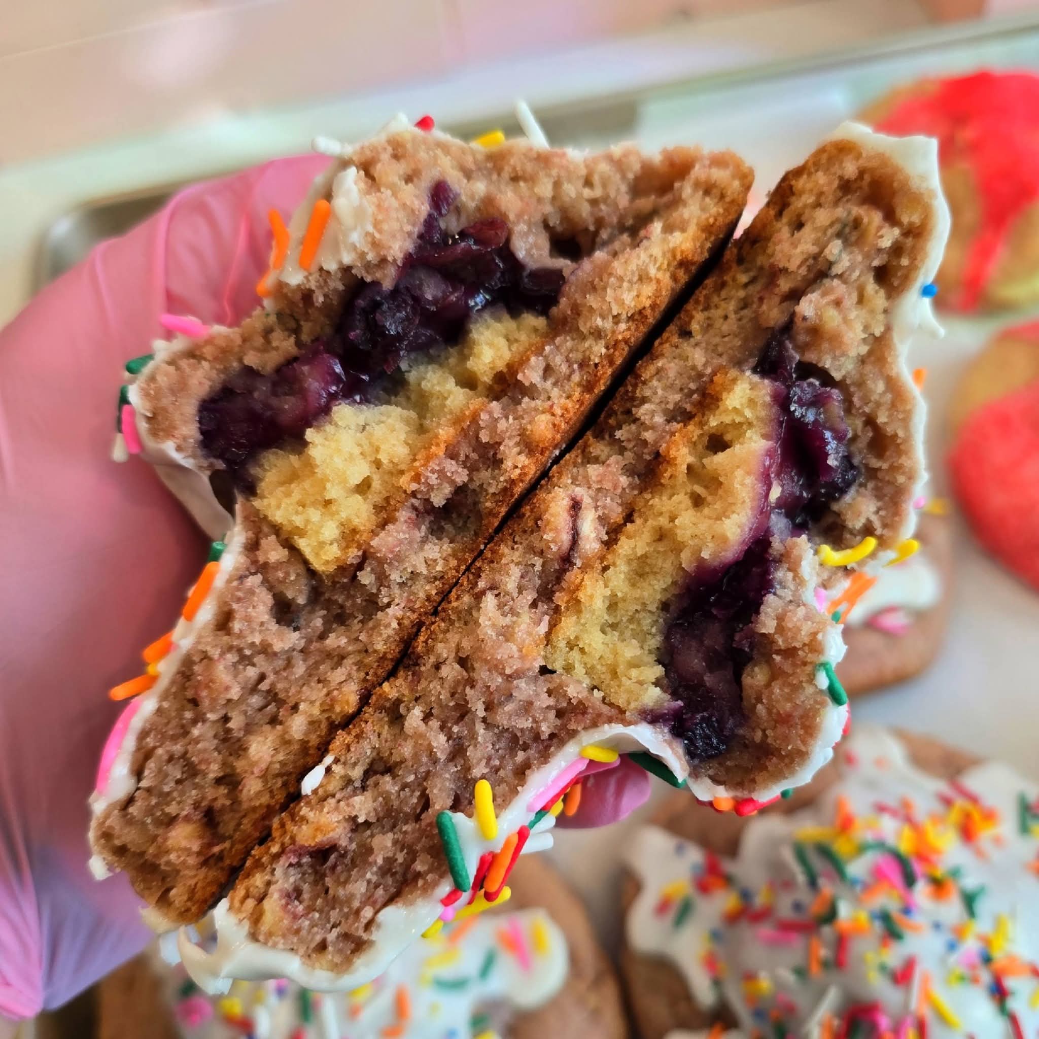 Close-up of a sliced cupcake with blueberry filling and colorful sprinkles, held in a hand with a pink glove.