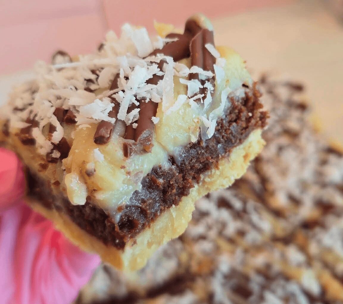Close-up of a layered dessert bar with chocolate, cream, and shredded coconut, held by a person with a pink glove.