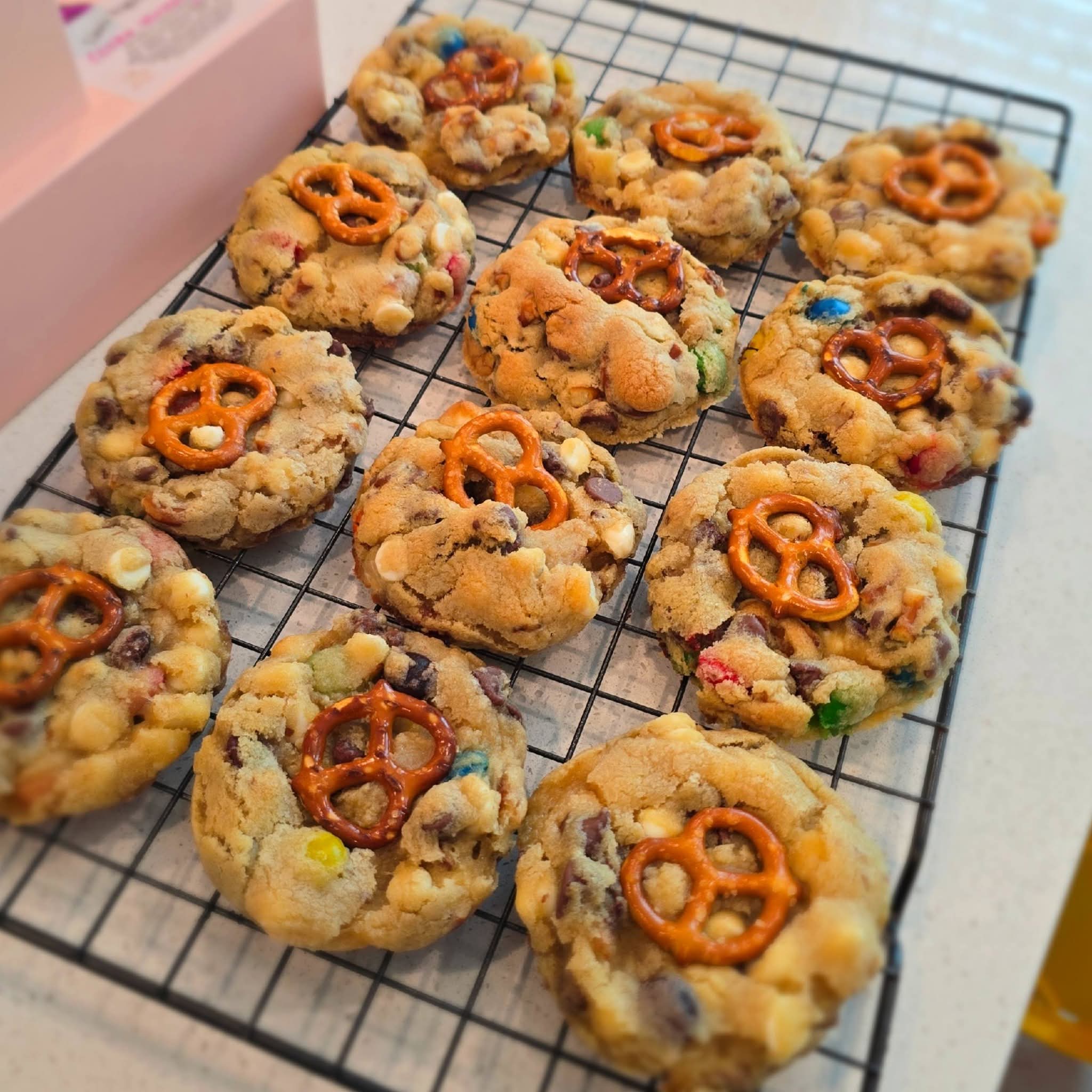 Cookies with pretzels and colorful candies on a cooling rack, in a pink box nearby.