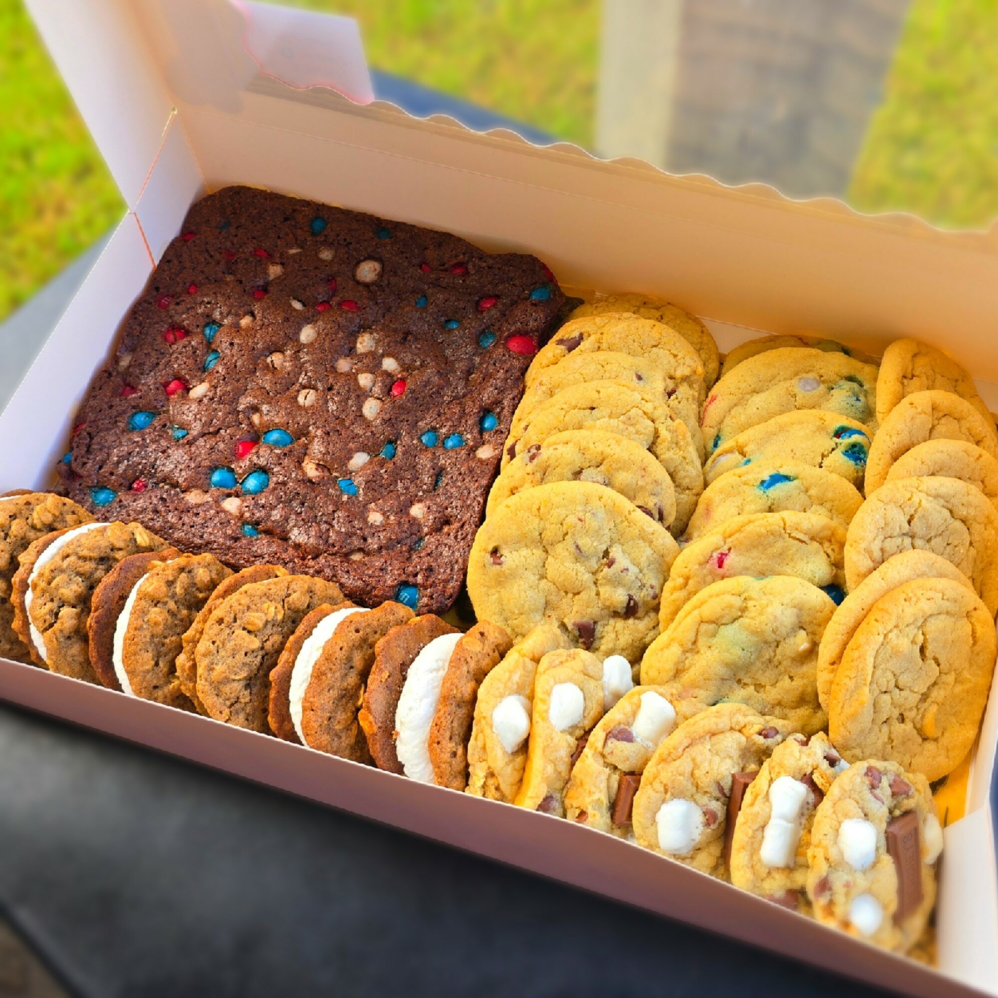 Box of assorted cookies with chocolate, colorful sprinkles, and white icing decorations, on a black surface with a blurred outdoor background.