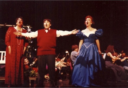 Three performers singing on stage with an orchestra in the background, arms outstretched, dressed in formal attire.