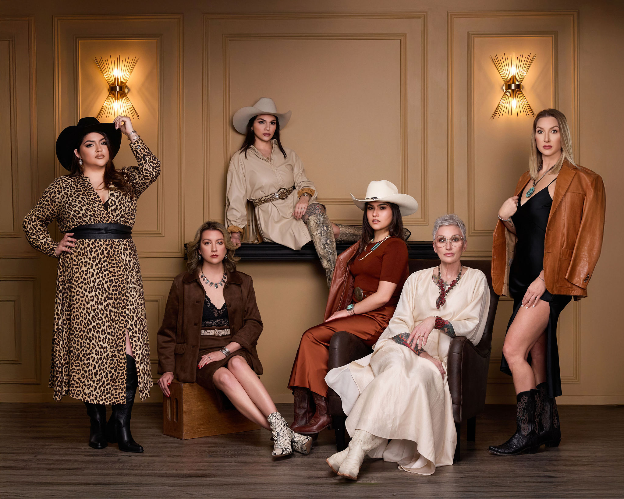 Six women in vintage and modern clothing pose in a decorated room with wall sconces and wooden paneling.