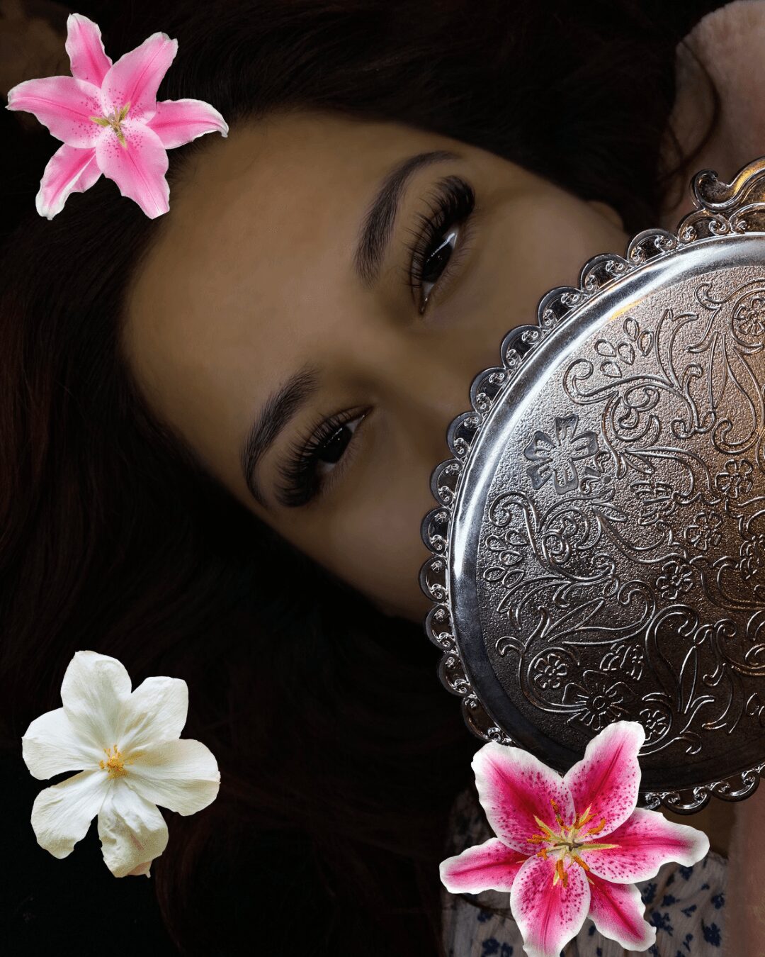 Woman with dark hair and makeup partially hidden behind a decorative silver mirror, surrounded by pink and white flowers.