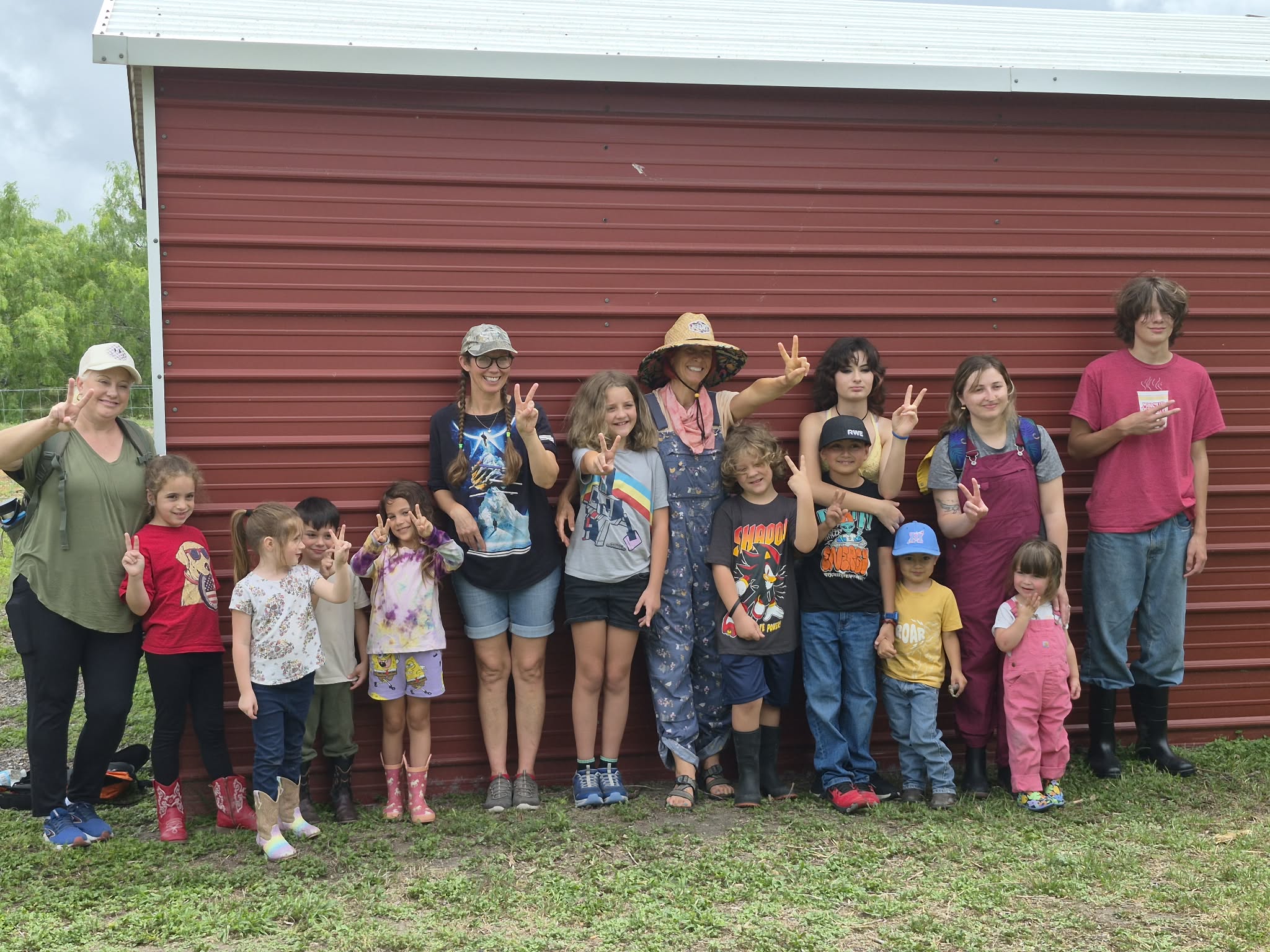Group of children and adults standing outdoors in front of a red building, some making peace signs, on grass.