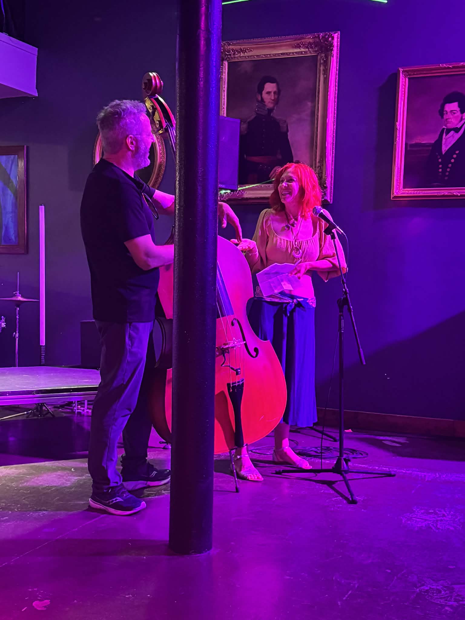 Two people on stage with a microphone and a double bass, in a room with framed portraits, purple lighting, and a pole.