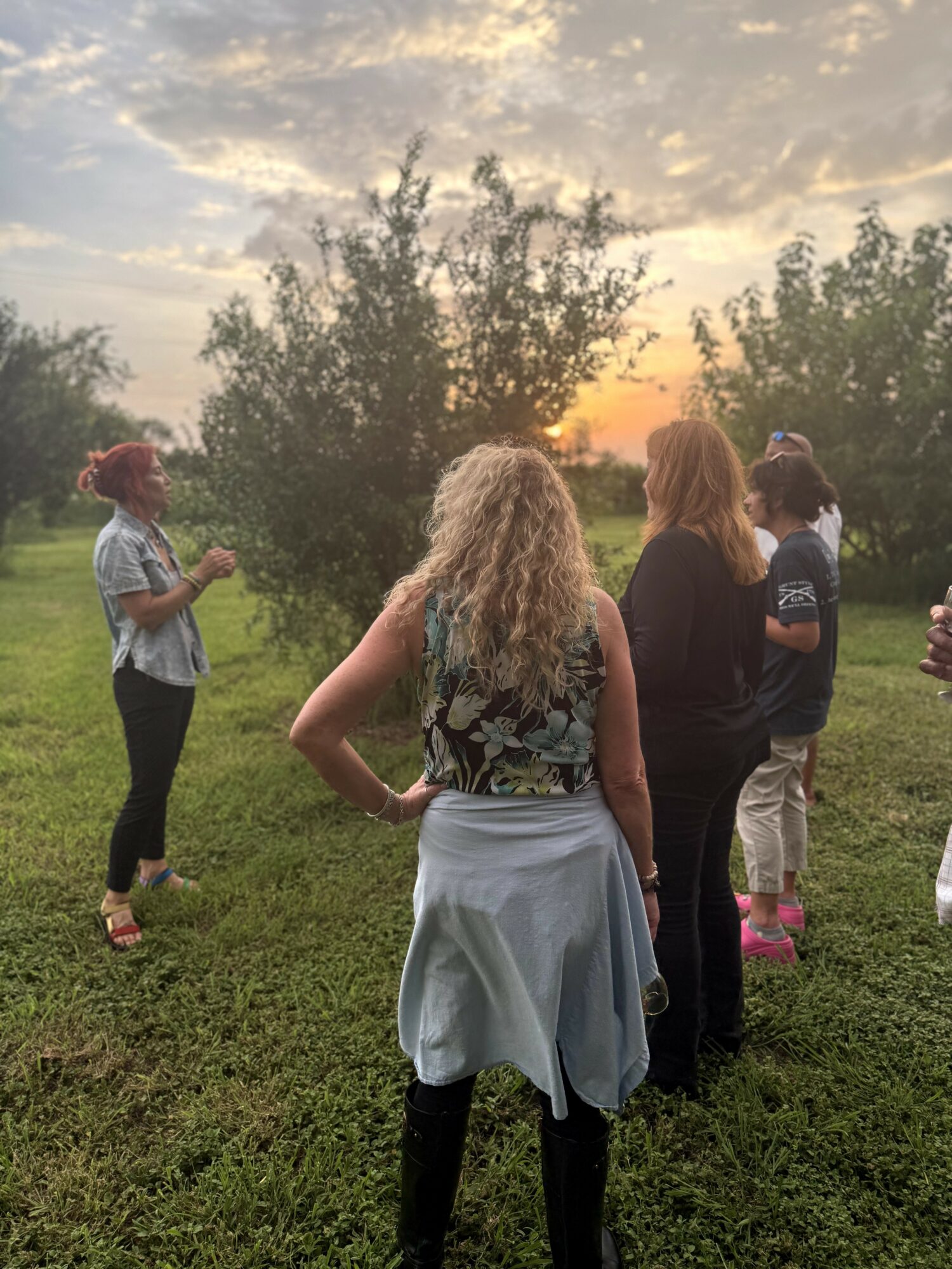 Group of people standing outdoors on grass during sunset, with trees and sky in background.