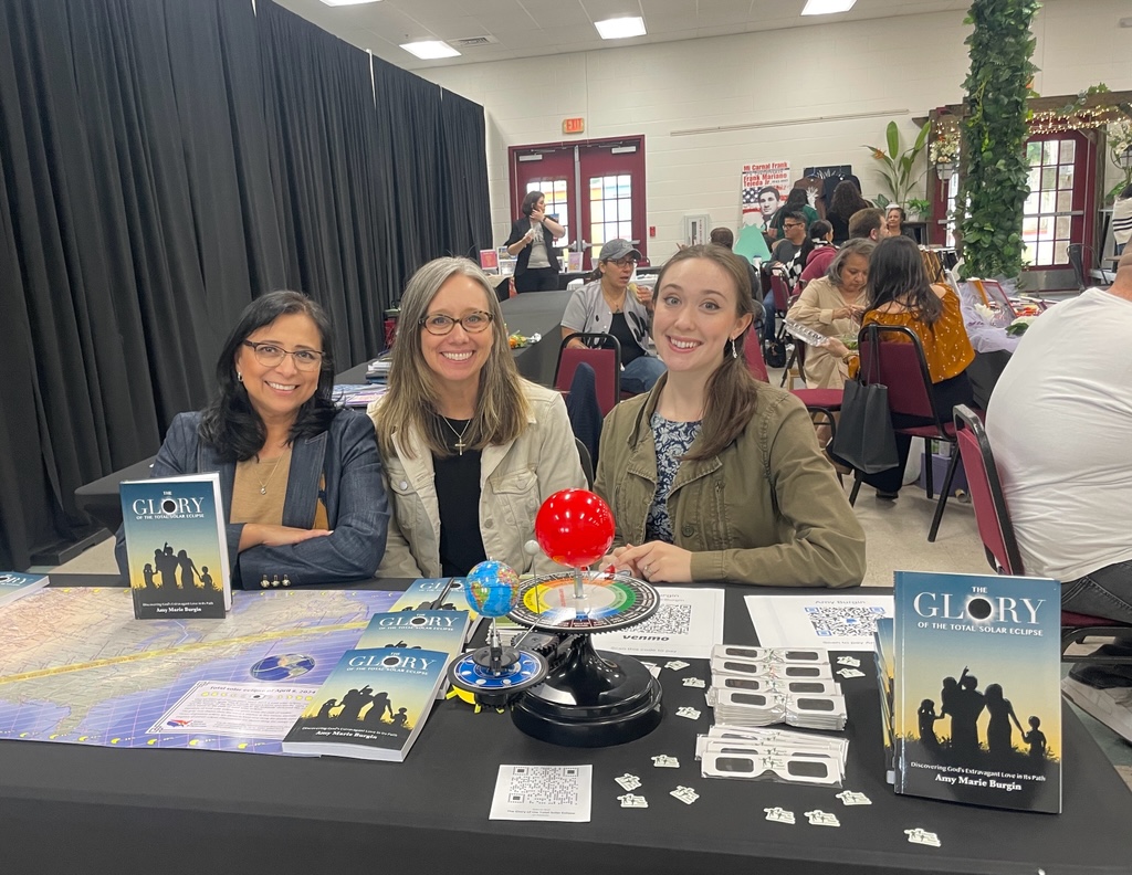 Three women sit at a table with board game materials, smiling, in a busy indoor event space.