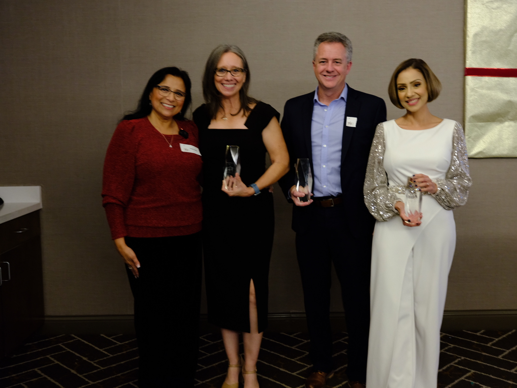 Four people standing together indoors, smiling, with two women holding awards or trophies.