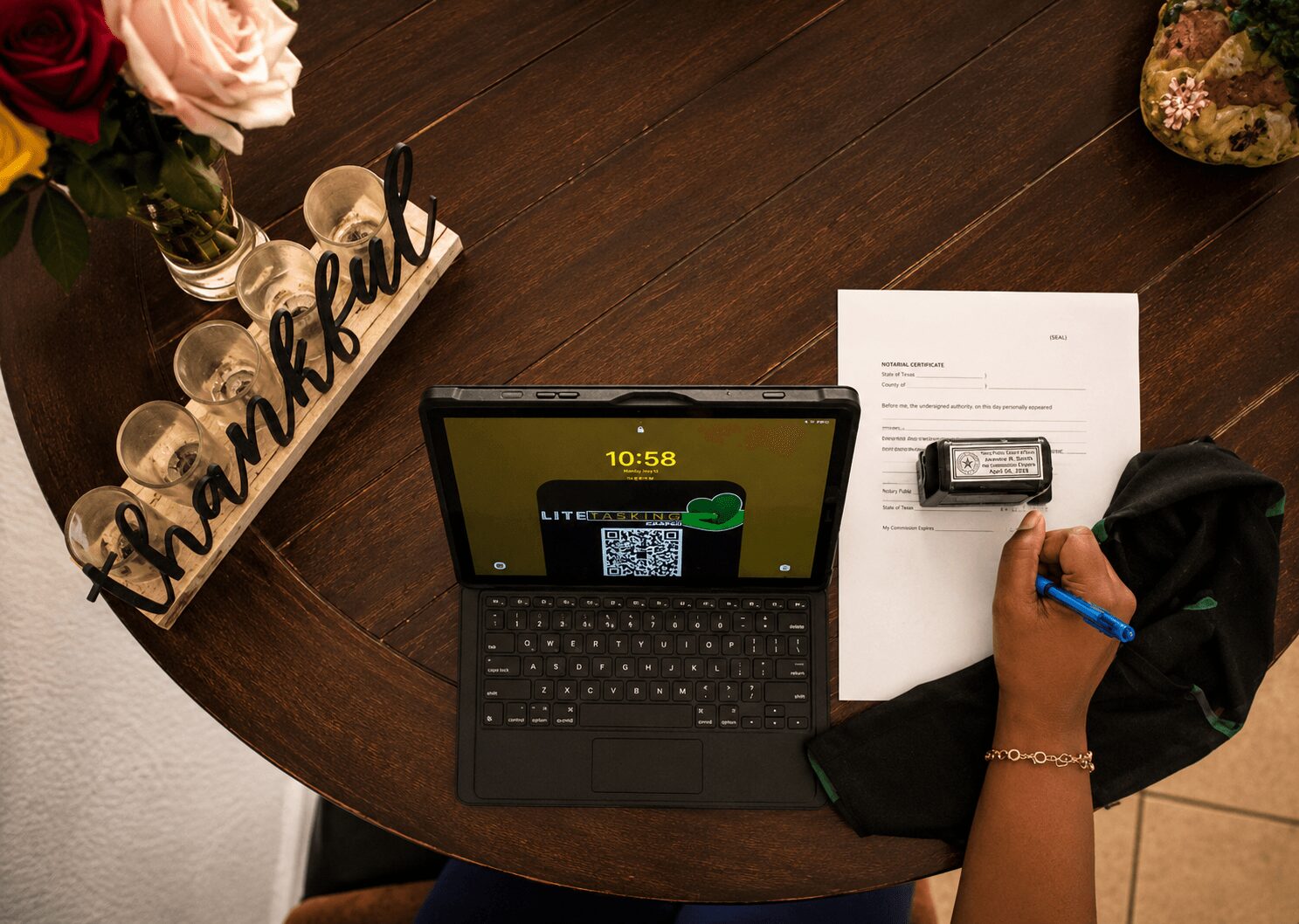 Person writing on paper with a pen, tablet, flowers, and a decorative 'thankful' sign on a wooden table.