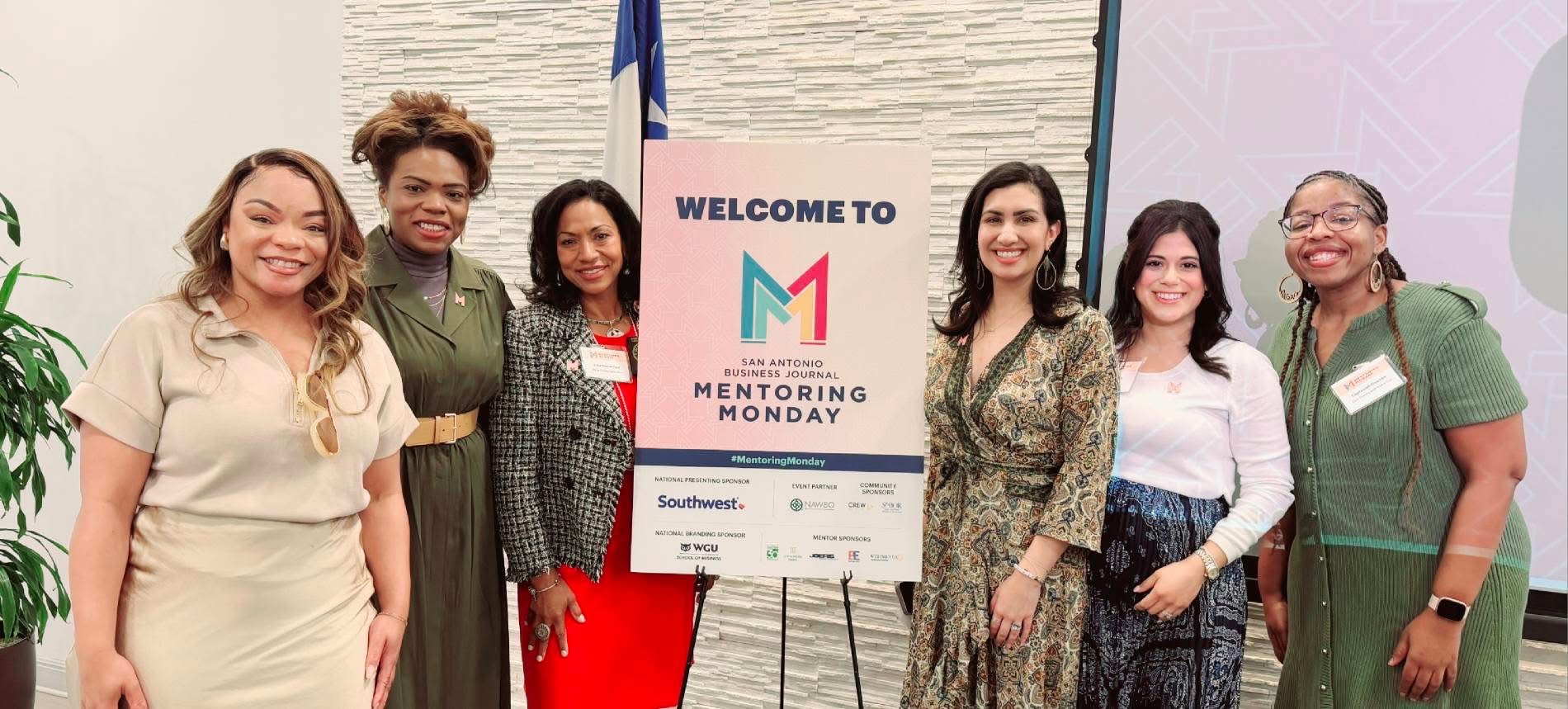 Six women standing together in front of a screen and a sign that reads 'Welcome to Mentoring Monday.'