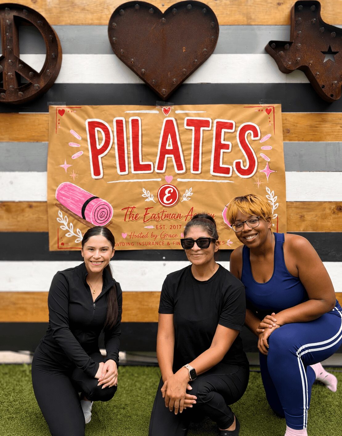 Three women sitting in front of a Pilates sign, smiling, with decorative hearts and peace sign on a wooden background.