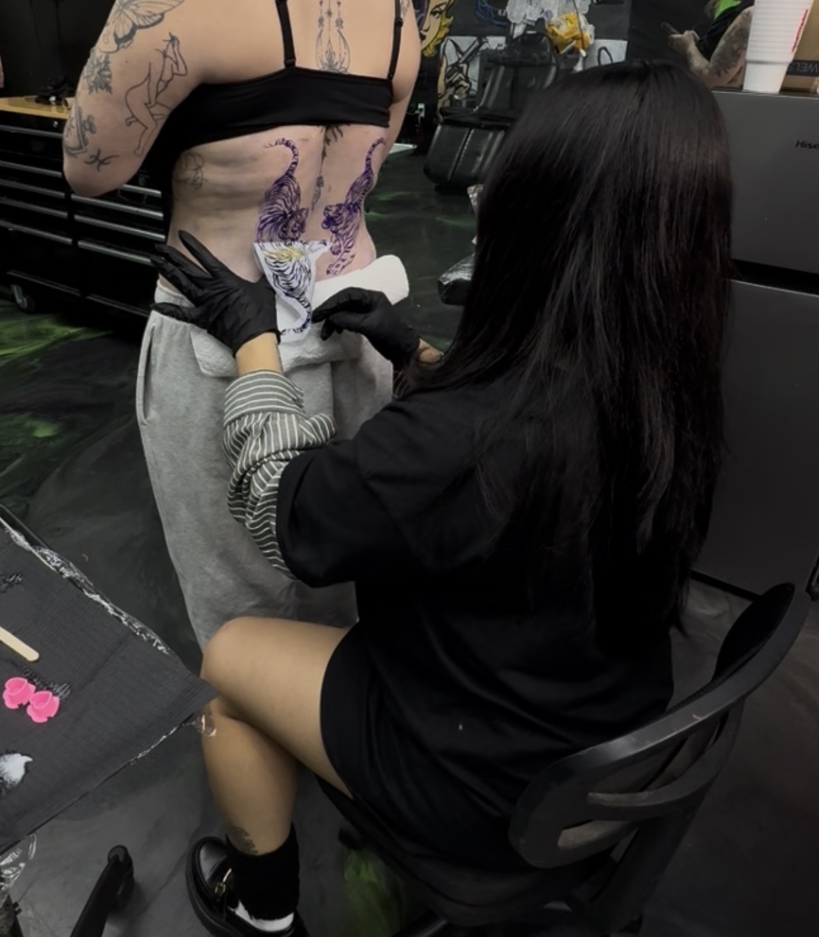 Tattooed person with exposed midriff and a tattooed arm, seated while a woman in black clothes and gloves tattoos their side.