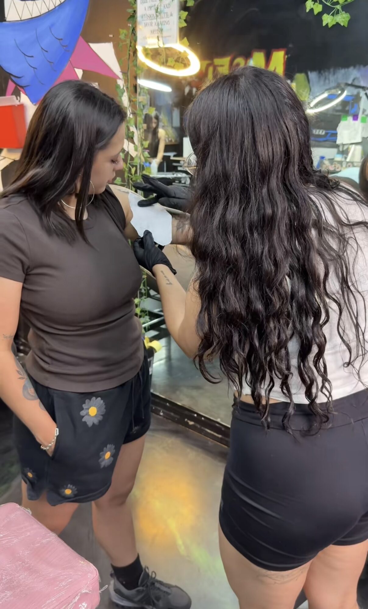 Two women are in a store, one is getting a tattoo on her arm, the other is assisting. They are near a glass window.
