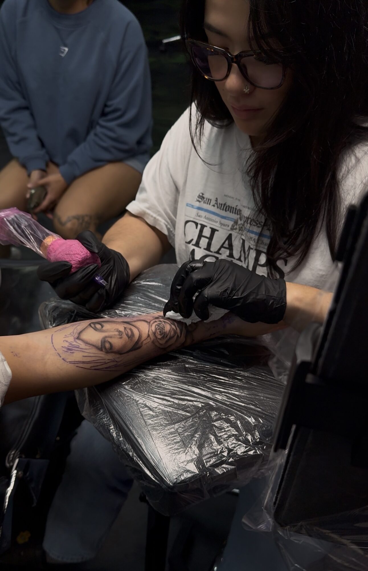 Person getting a tattoo on their arm by a tattoo artist wearing black gloves, with another person nearby. Dark background.