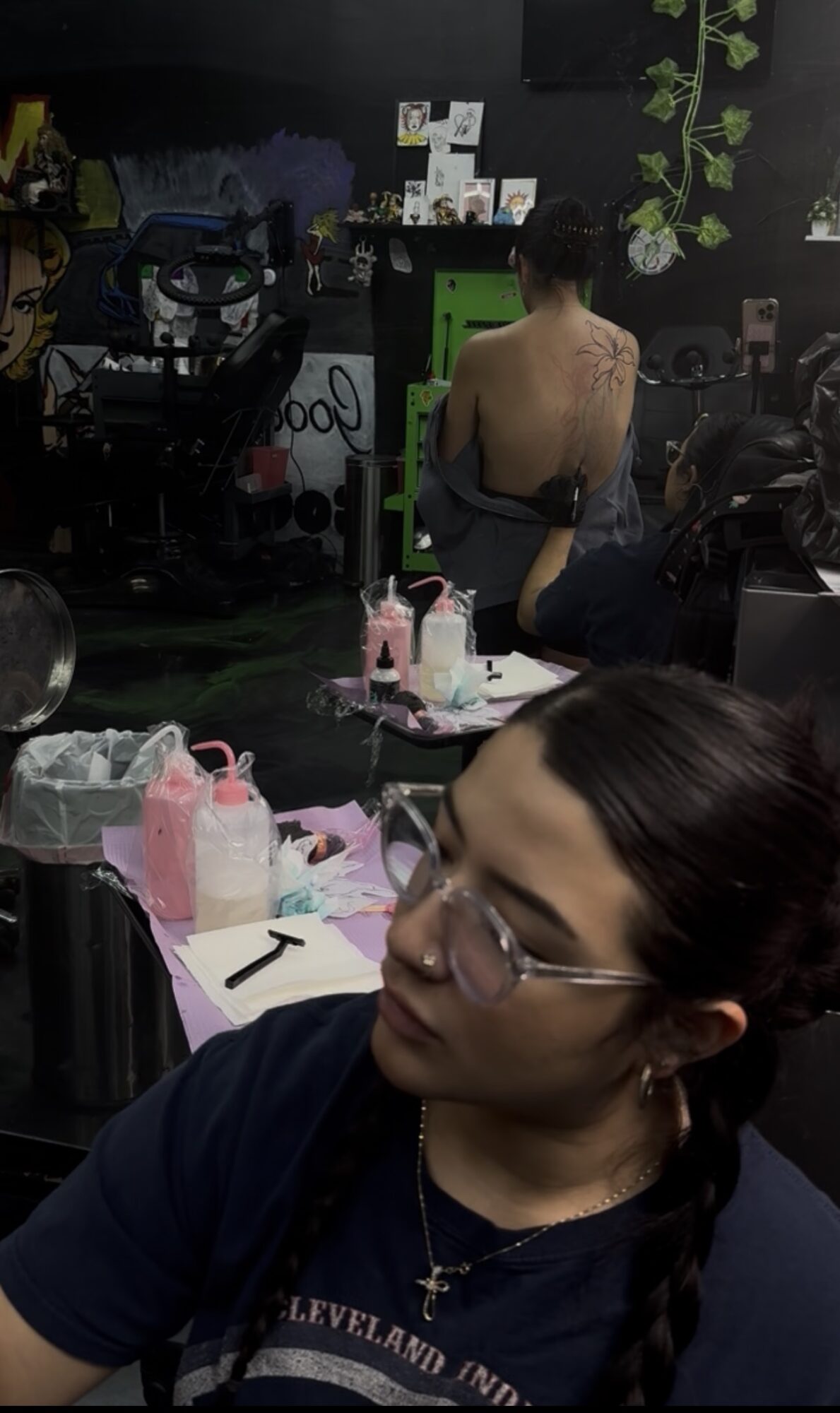 Woman with glasses and braided hair sitting at a table with pink and white items, in a room with a person in the background.