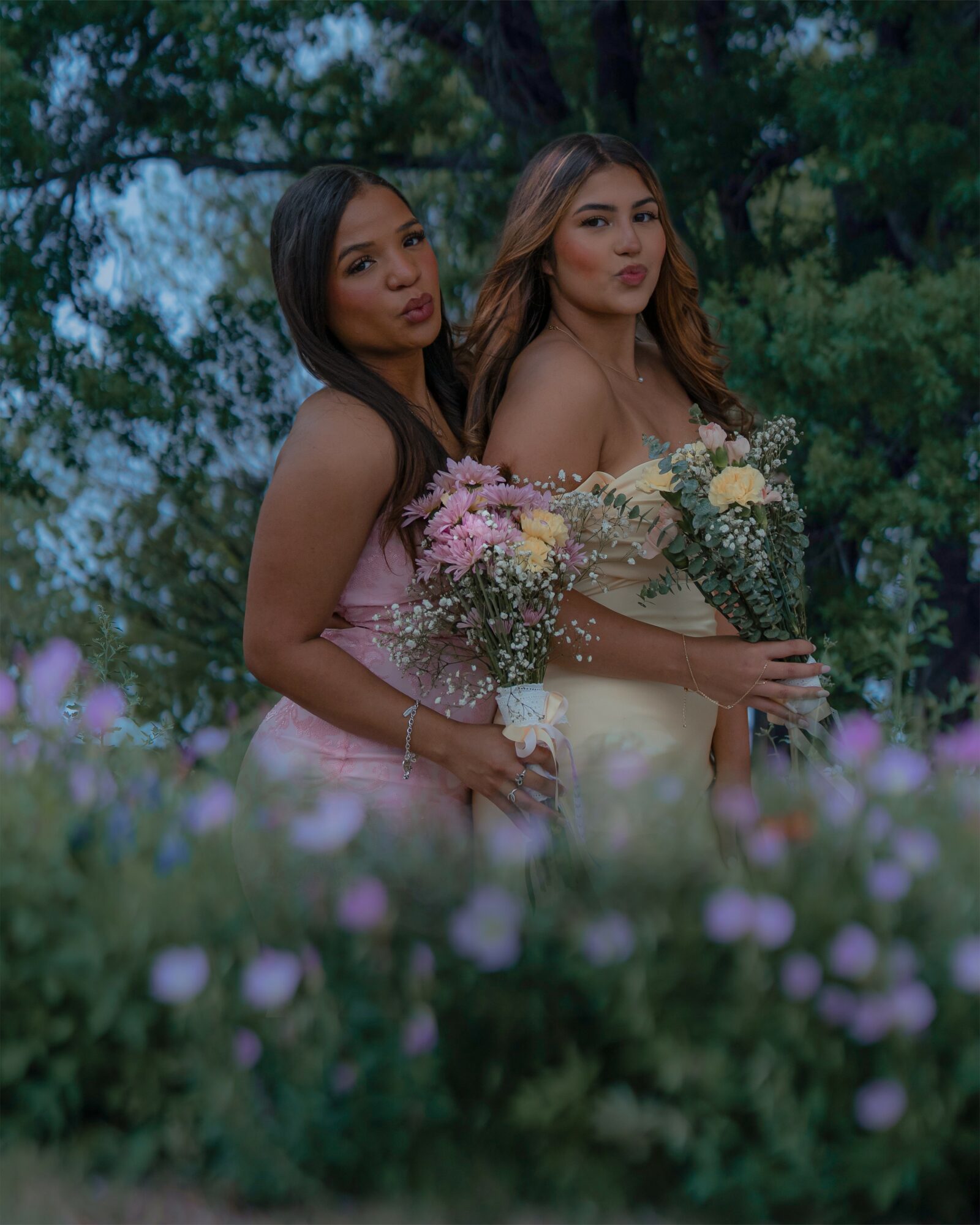 Two women holding bouquets of flowers outdoors with trees in the background, surrounded by purple flowers in the foreground.
