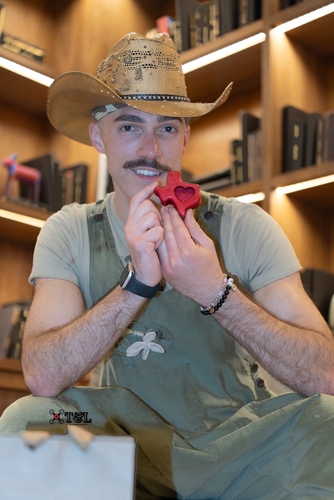 Person wearing a cowboy hat and apron, holding a red heart-shaped object, smiling in a library setting.