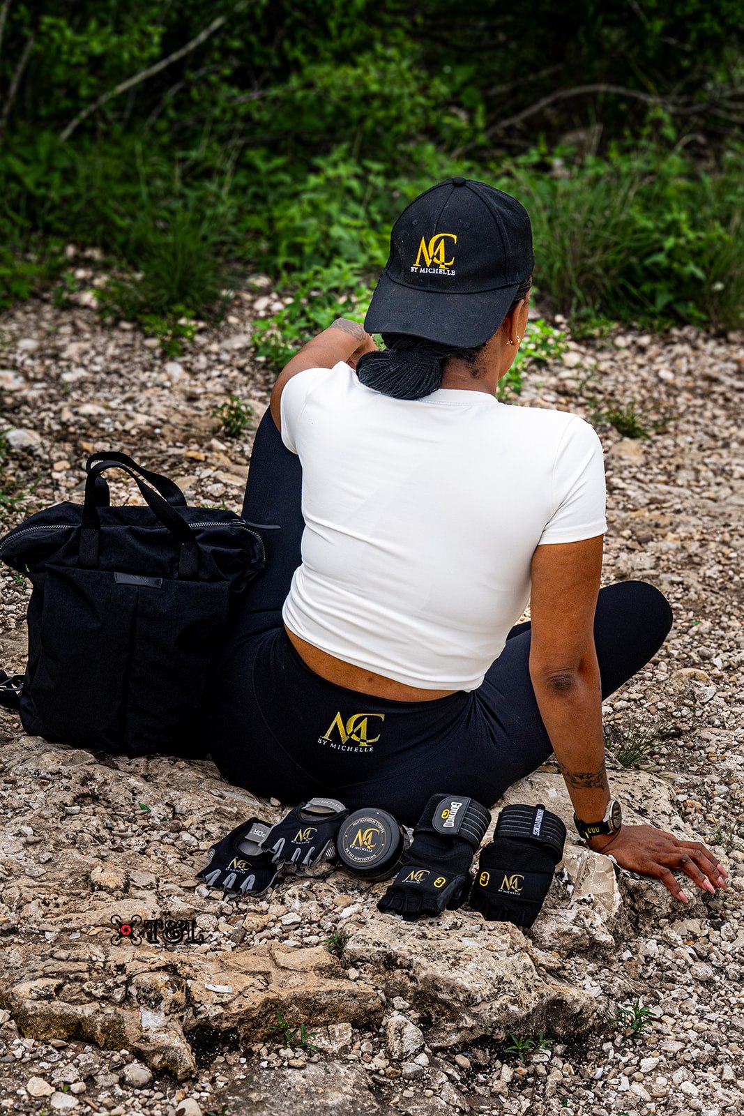 Person sitting on rocky ground with a black bag and equipment, wearing a white shirt, black pants, and a cap, outdoors.