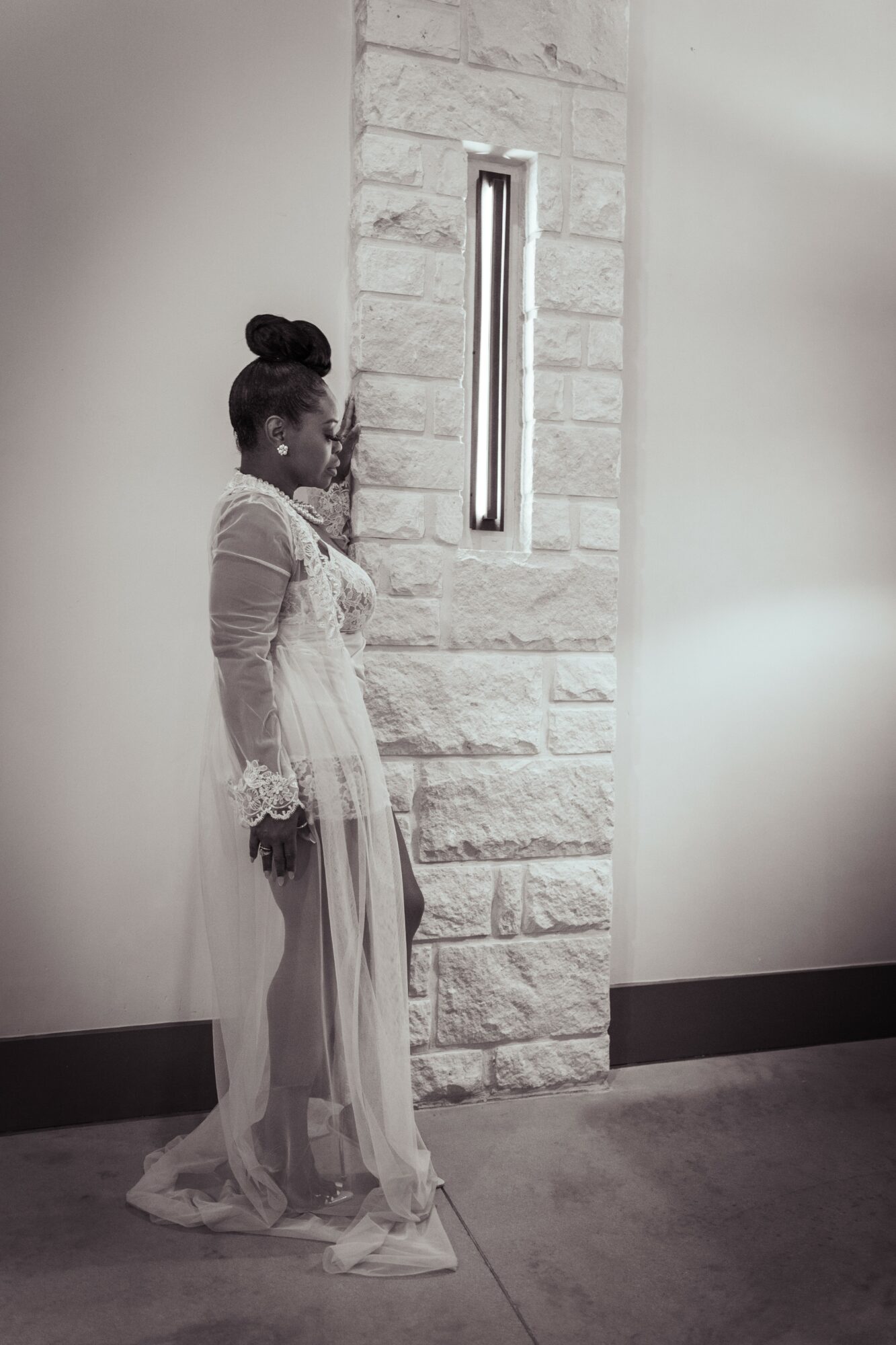 Woman in a long dress standing against a stone wall with a vertical light fixture, looking to the side.