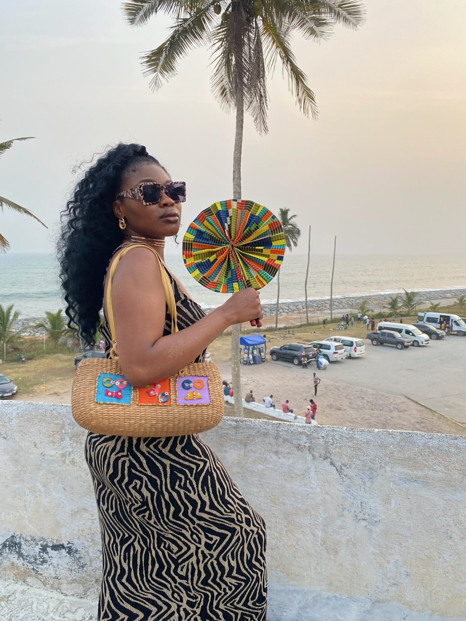Woman with sunglasses holding a colorful fan, standing outdoors near a beach with palm trees and parked cars.
