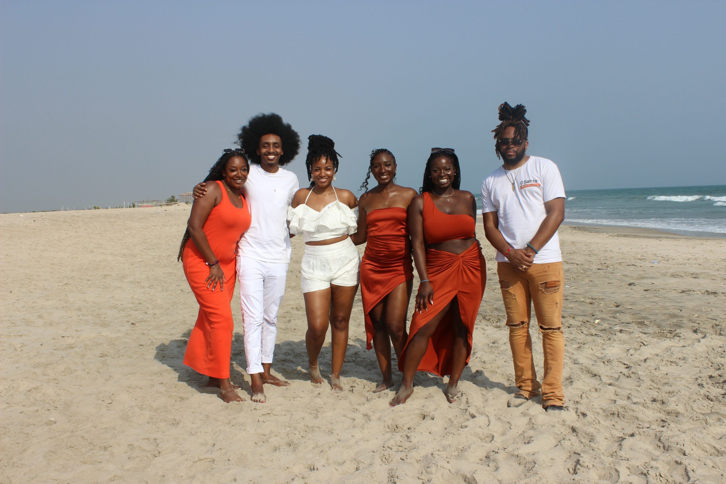 Six people standing on a sandy beach near the ocean, smiling and posing for a photo.